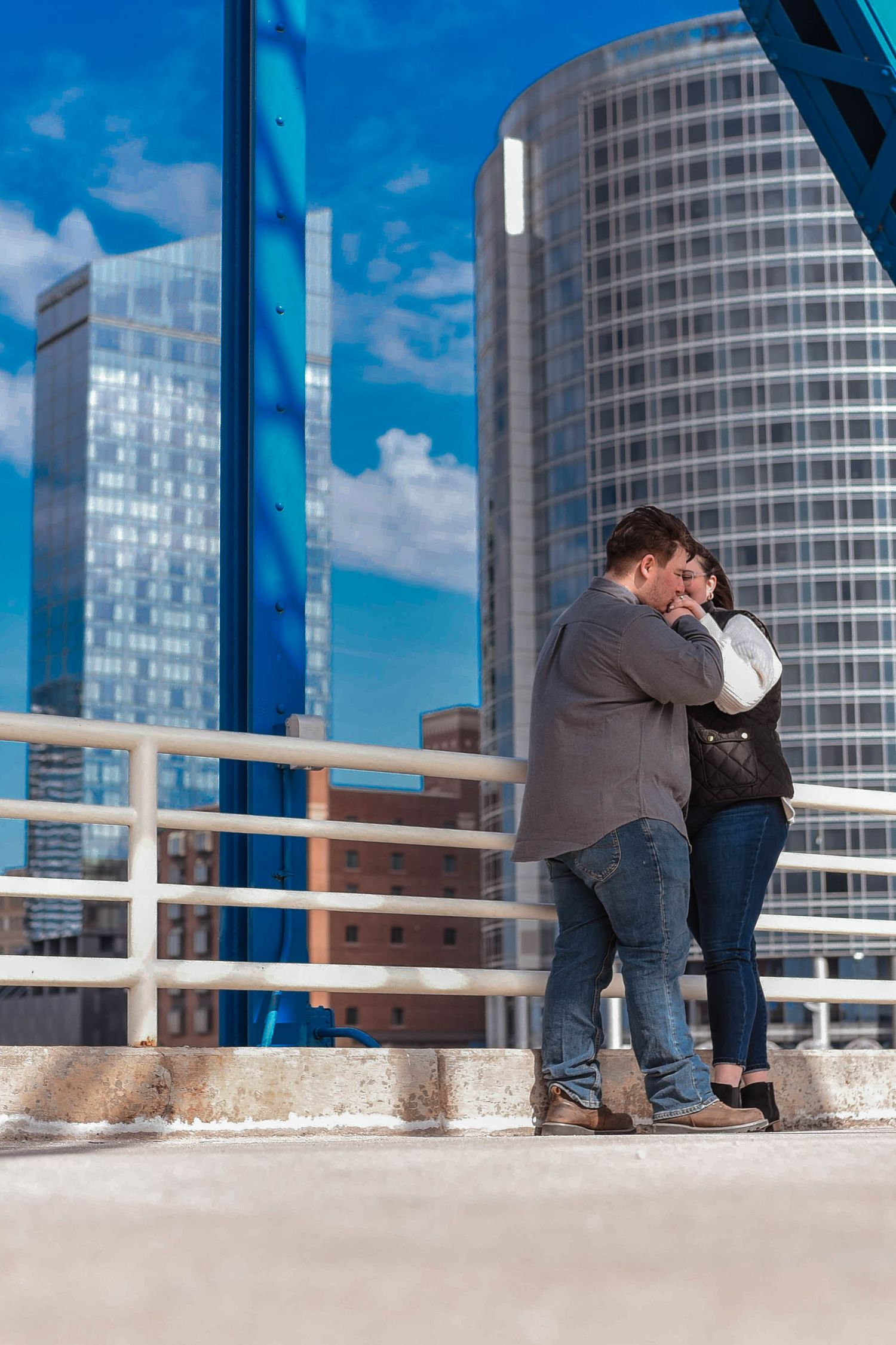 A couple shares a romantic moment on a bridge with modern buildings and blue sky in the background.