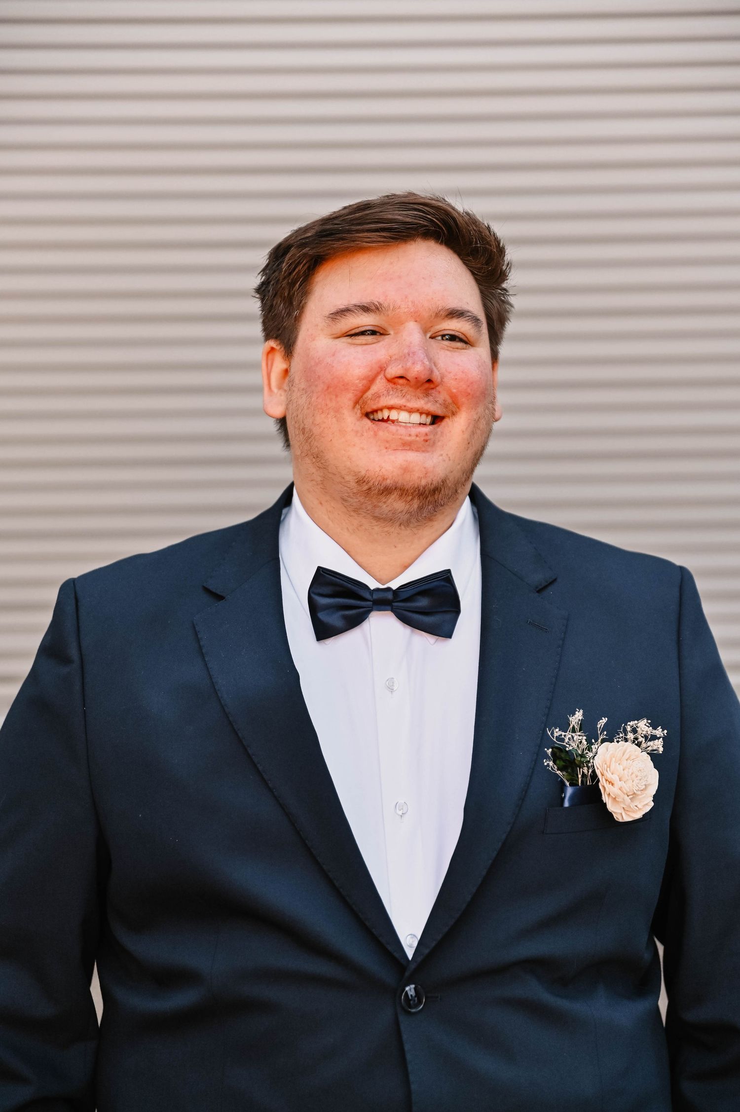 A groom in a navy suit and black bow tie smiles at the camera against a grey background.