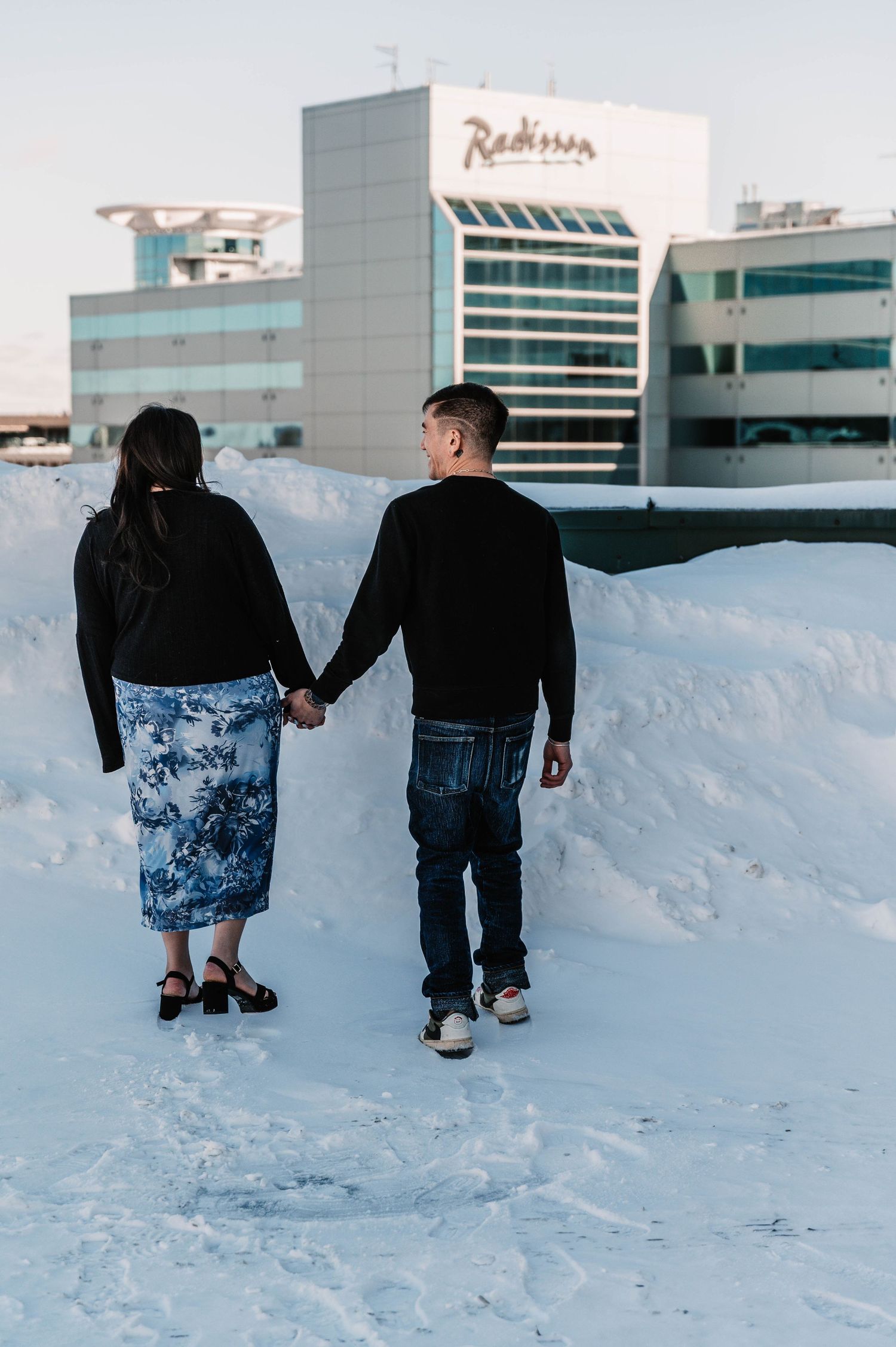 Couple walking hand in hand through snowy landscape with city buildings in background.