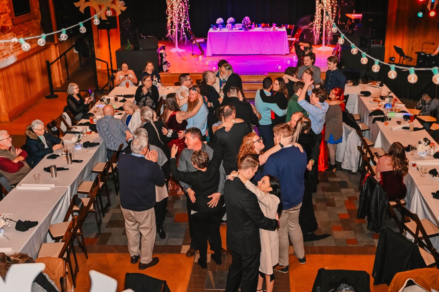 Large group of wedding guests mingling and dancing at indoor reception with orange ambient lighting.