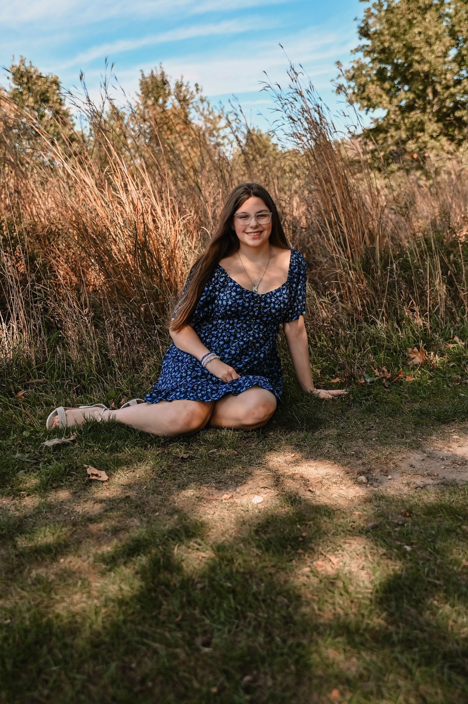 Person in a navy polka dot dress sits in a grassy area on a sunny day with trees in the background.