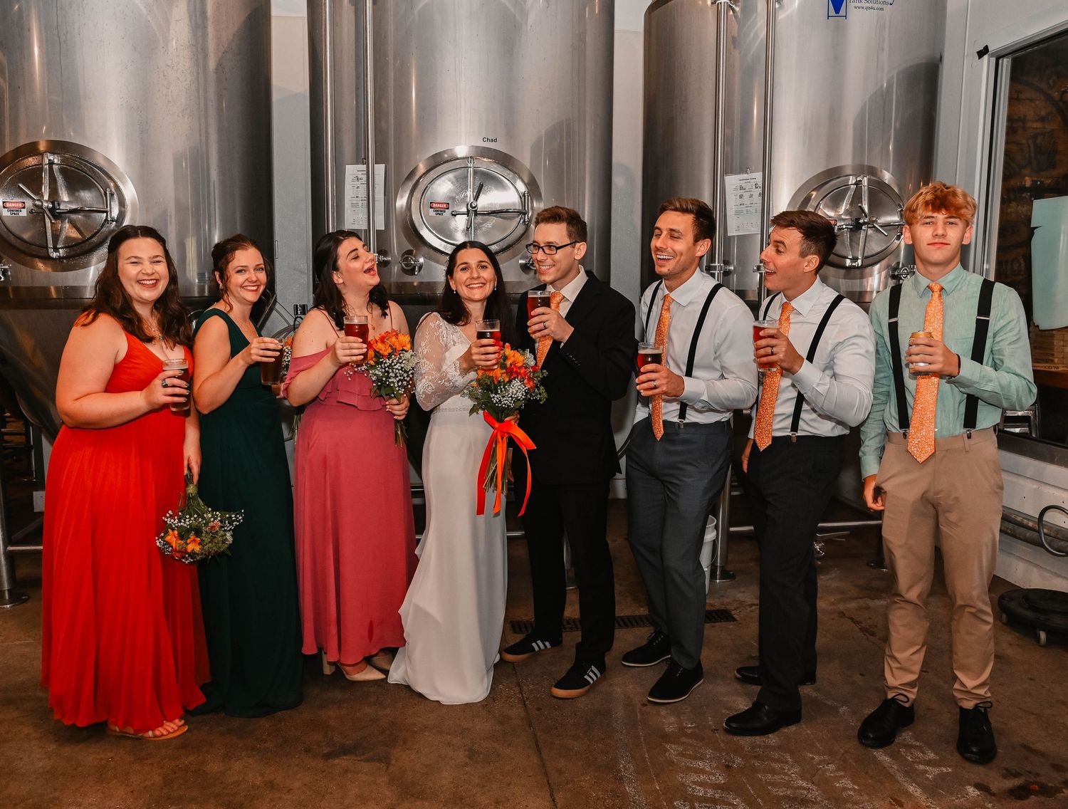 Wedding party members in red dresses and tan suits pose together for formal photos.