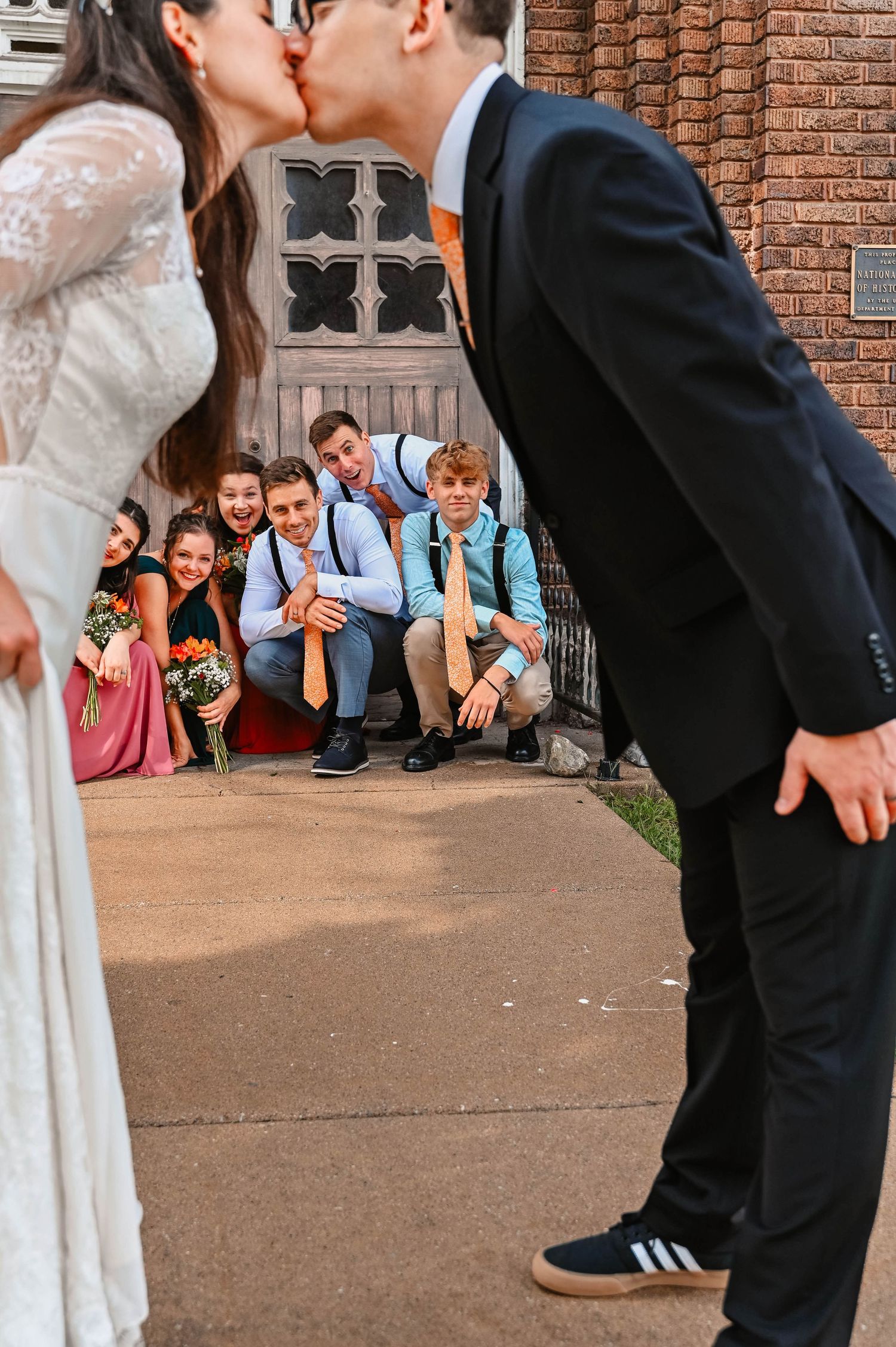 Wedding party peeks around corner during couple's kiss in front of brick church.