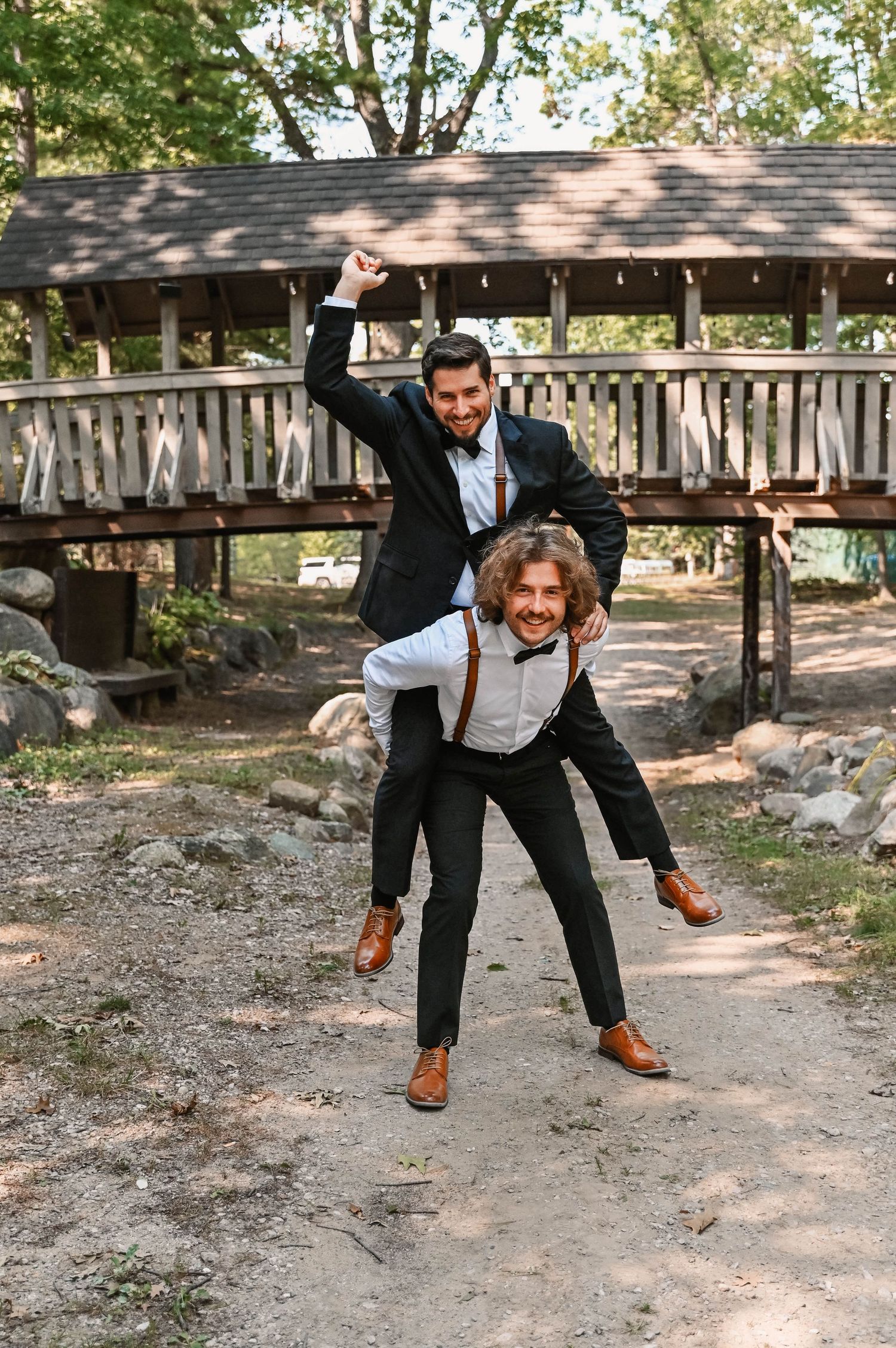 Playful jumping pose in front of rustic wooden bridge at outdoor venue.