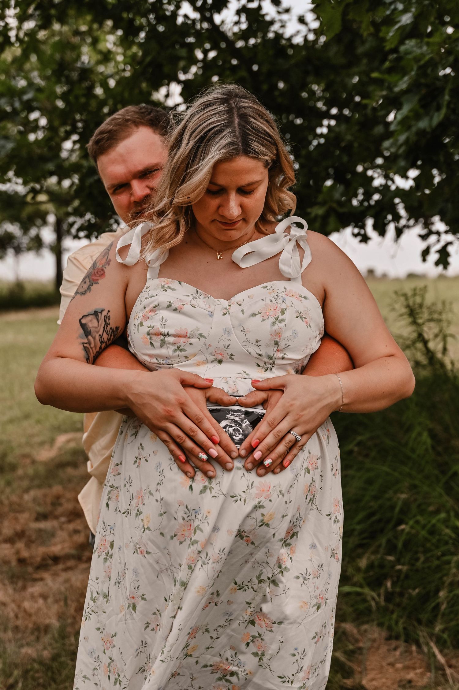A couple embraces while looking at an ultrasound photo in a floral dress outdoors.