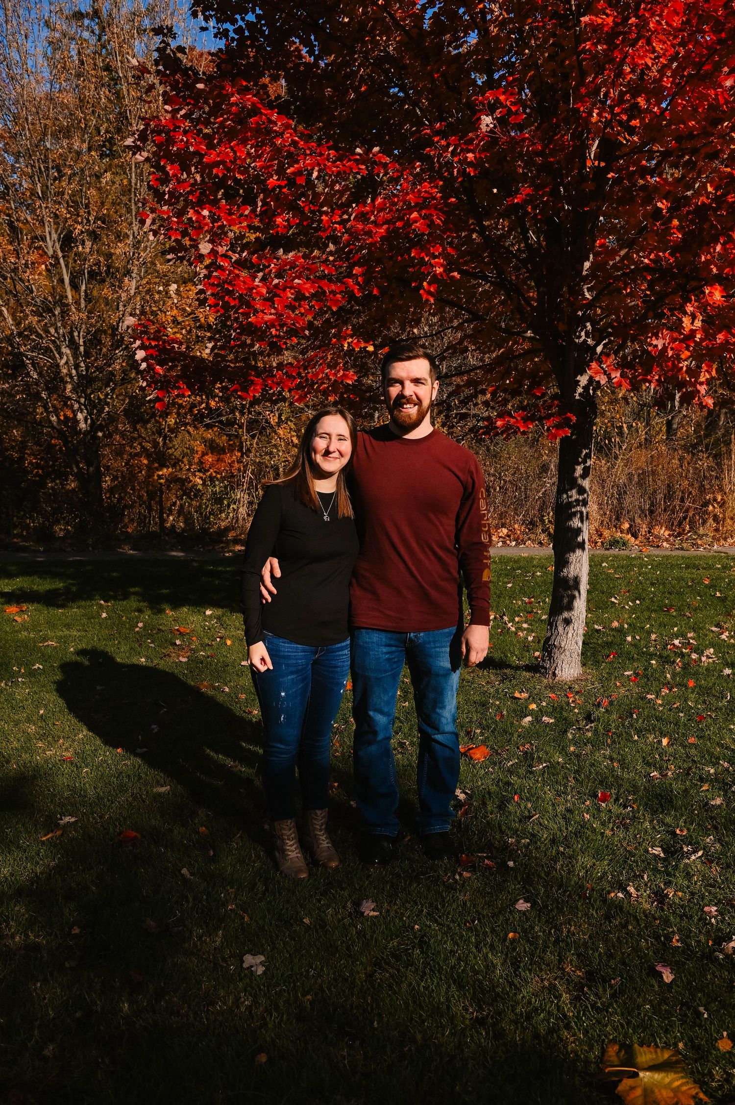 A couple poses together against vibrant red autumn foliage in a park setting.