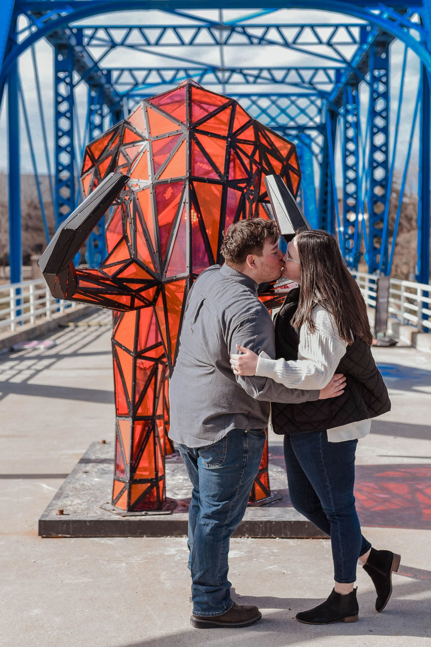 A couple embraces near orange geometric sculptures on a blue bridge walkway.