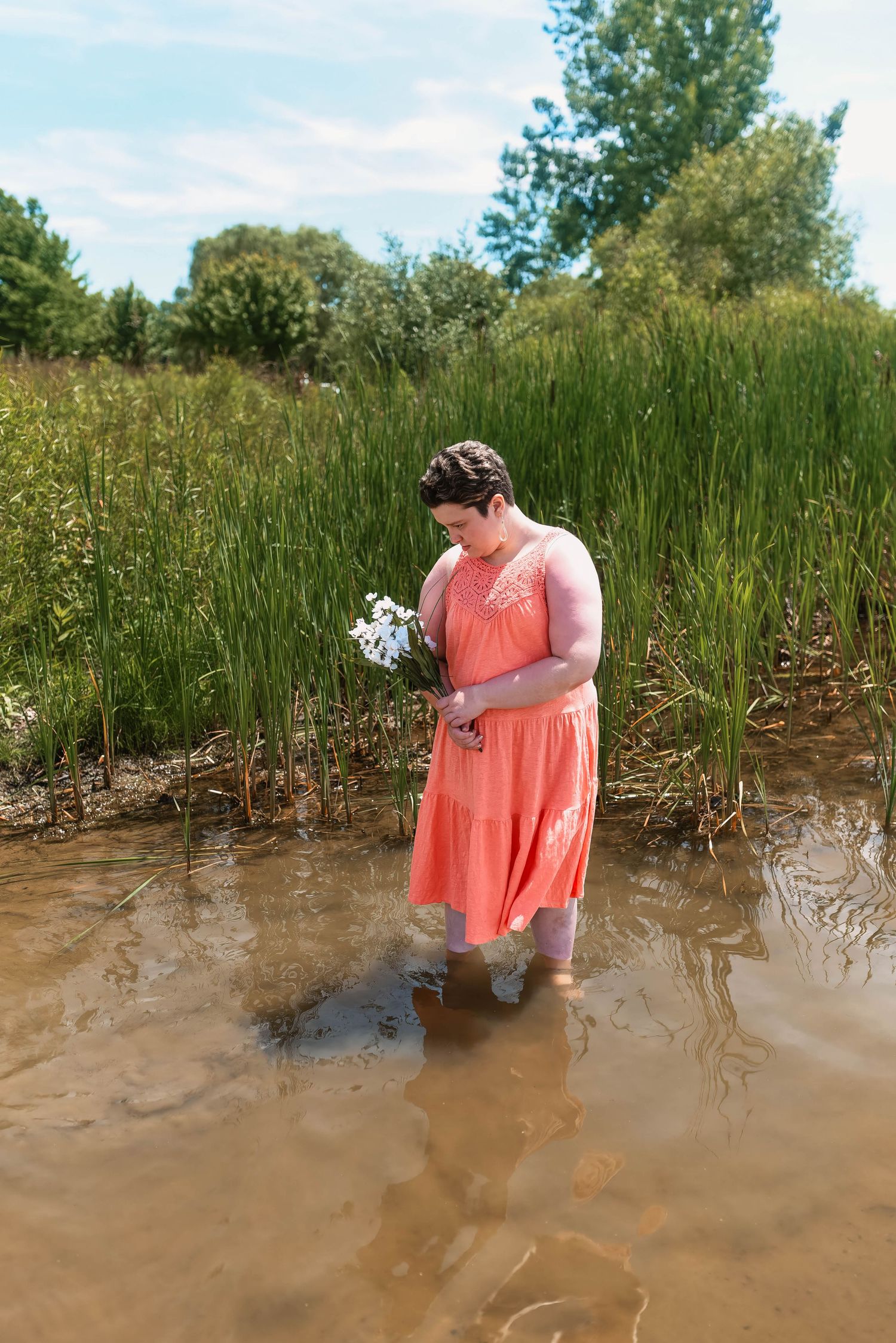 A person in a coral dress stands in muddy water holding wildflowers.
