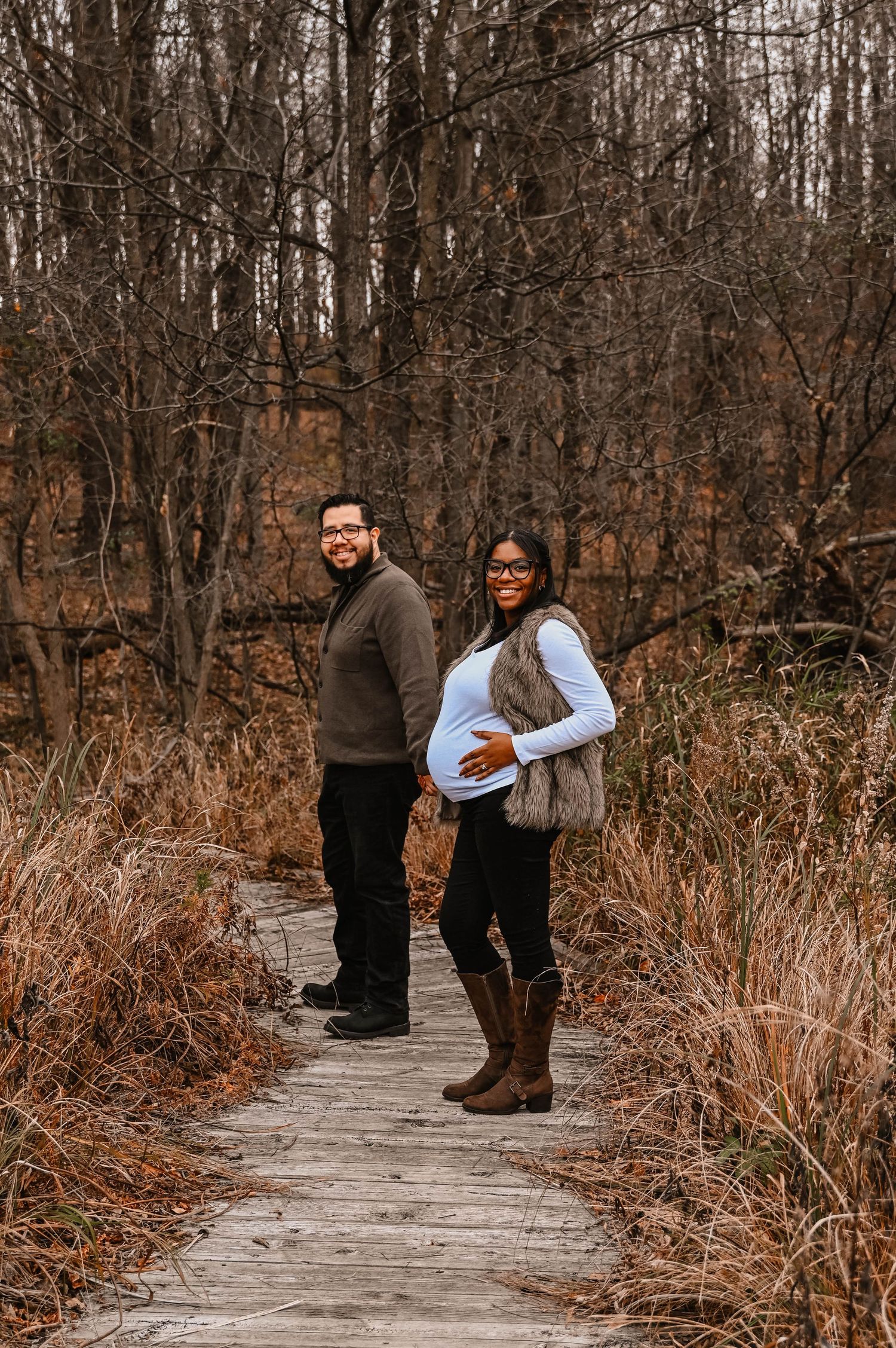 A couple walks along a wooden boardwalk surrounded by autumn grass and bare trees.