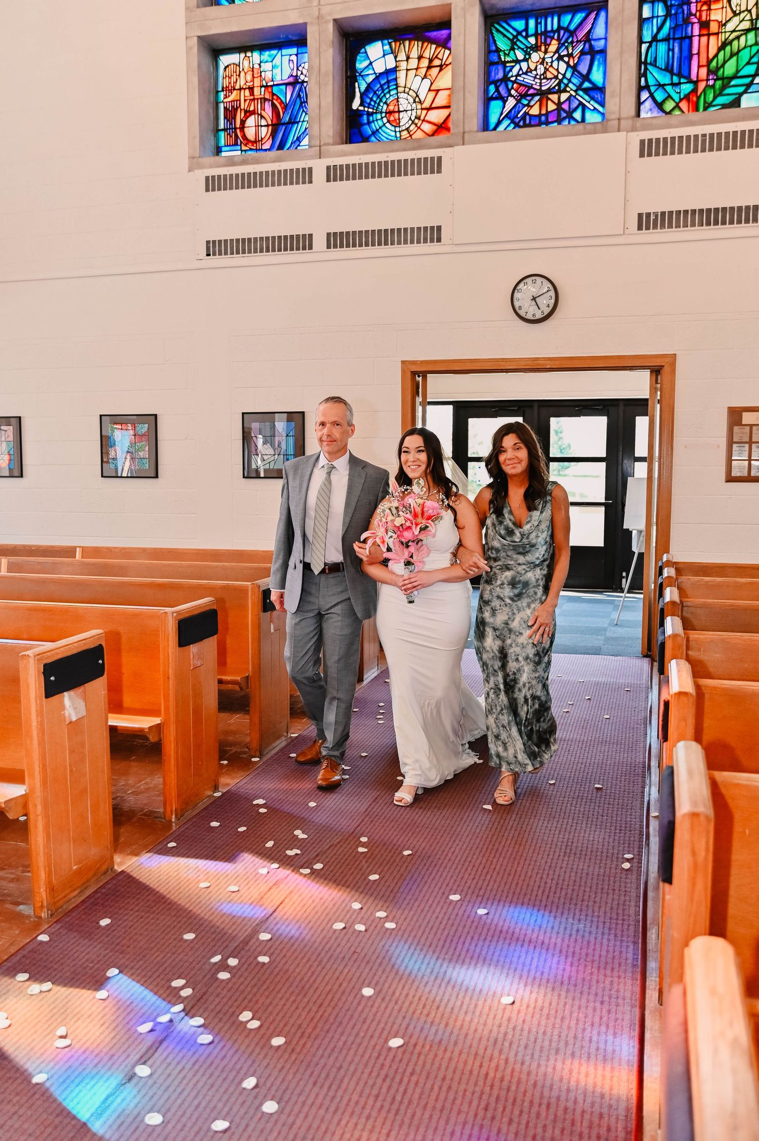 Wedding party walking down church aisle under stained glass windows with colorful light reflections.
