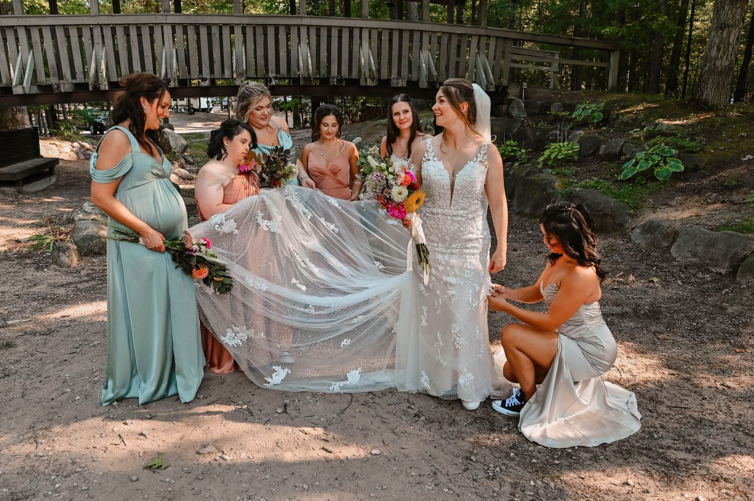 Bridesmaids in mint green dresses help arrange a wedding veil in an outdoor garden setting.