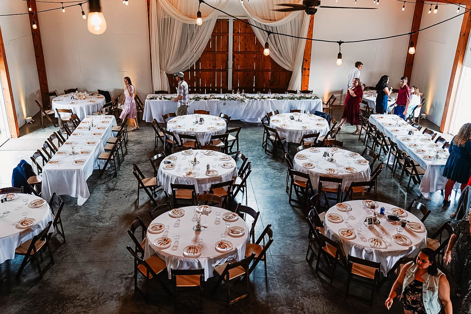 Reception venue set up with round tables, white linens and rustic decor in a barn setting.