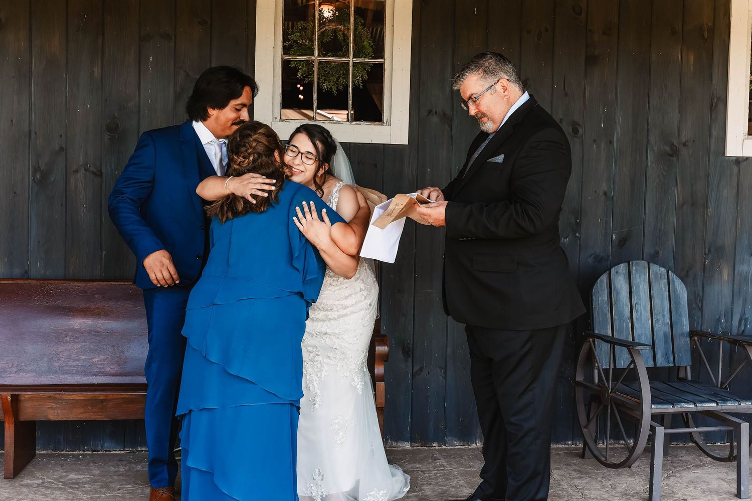 Wedding party poses outside a rustic dark wooden building with windows.