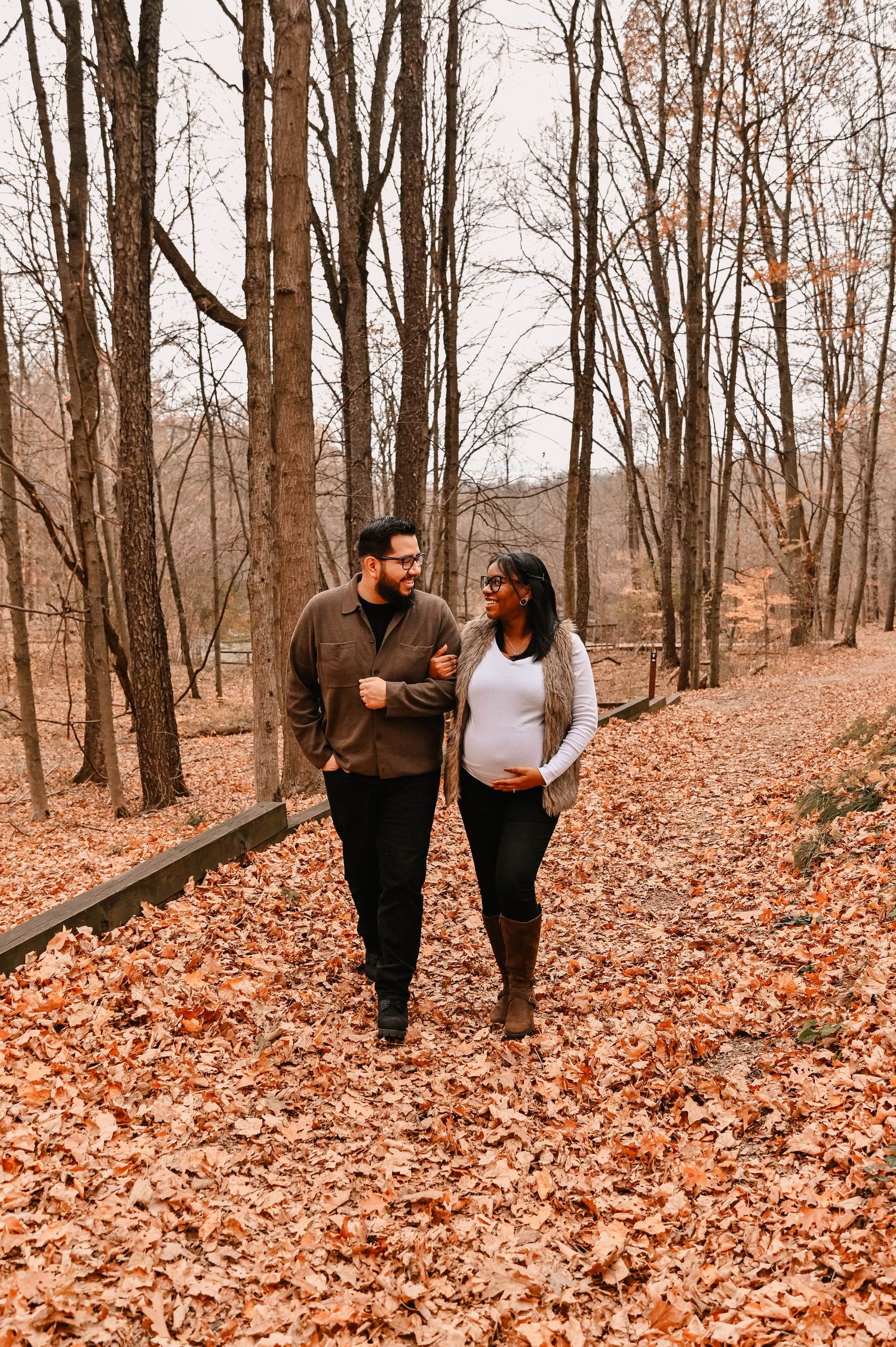 A couple walks hand in hand through a forest path covered in fallen autumn leaves.
