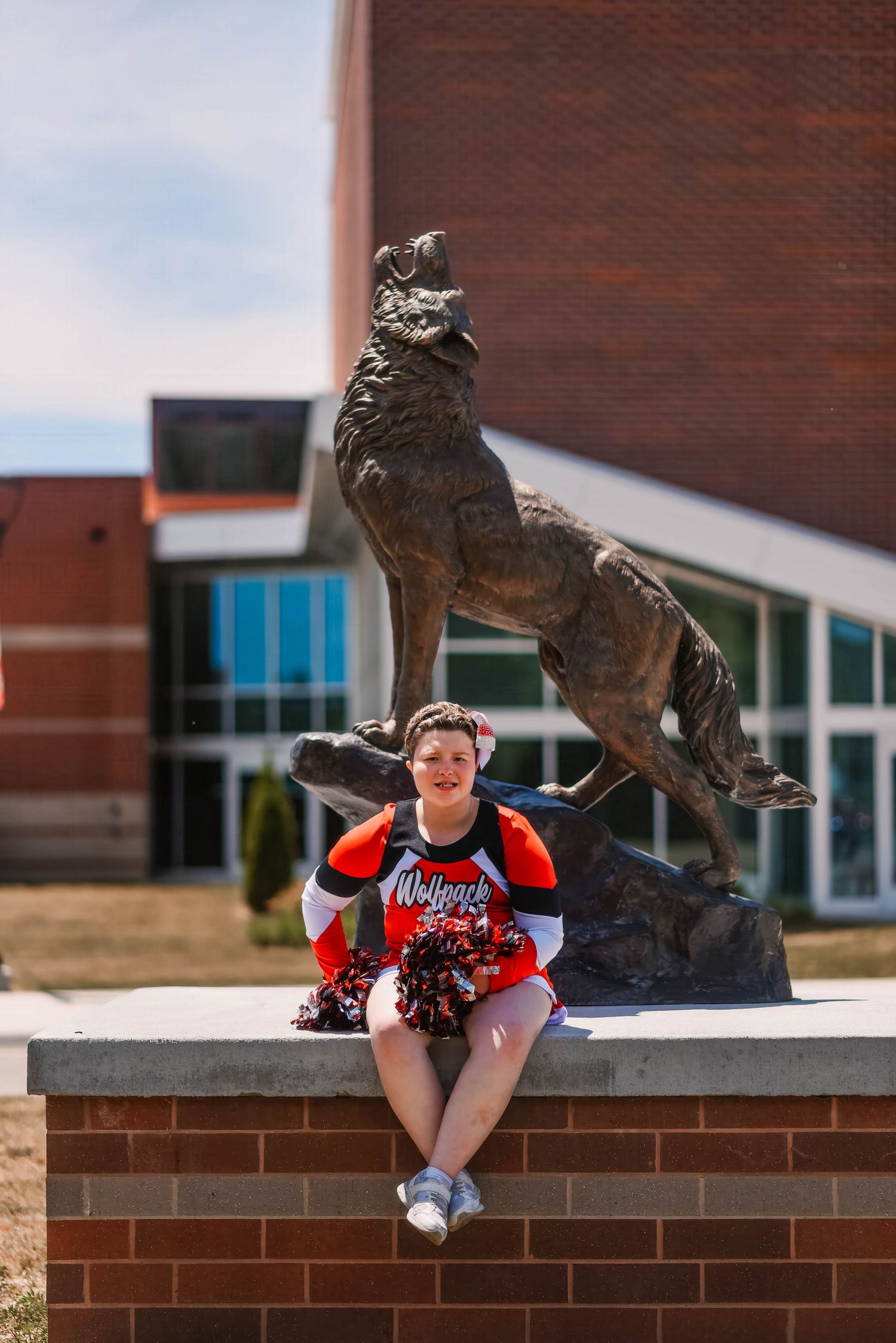 Cheerleader posing playfully by a bronze mascot statue outside a brick building.