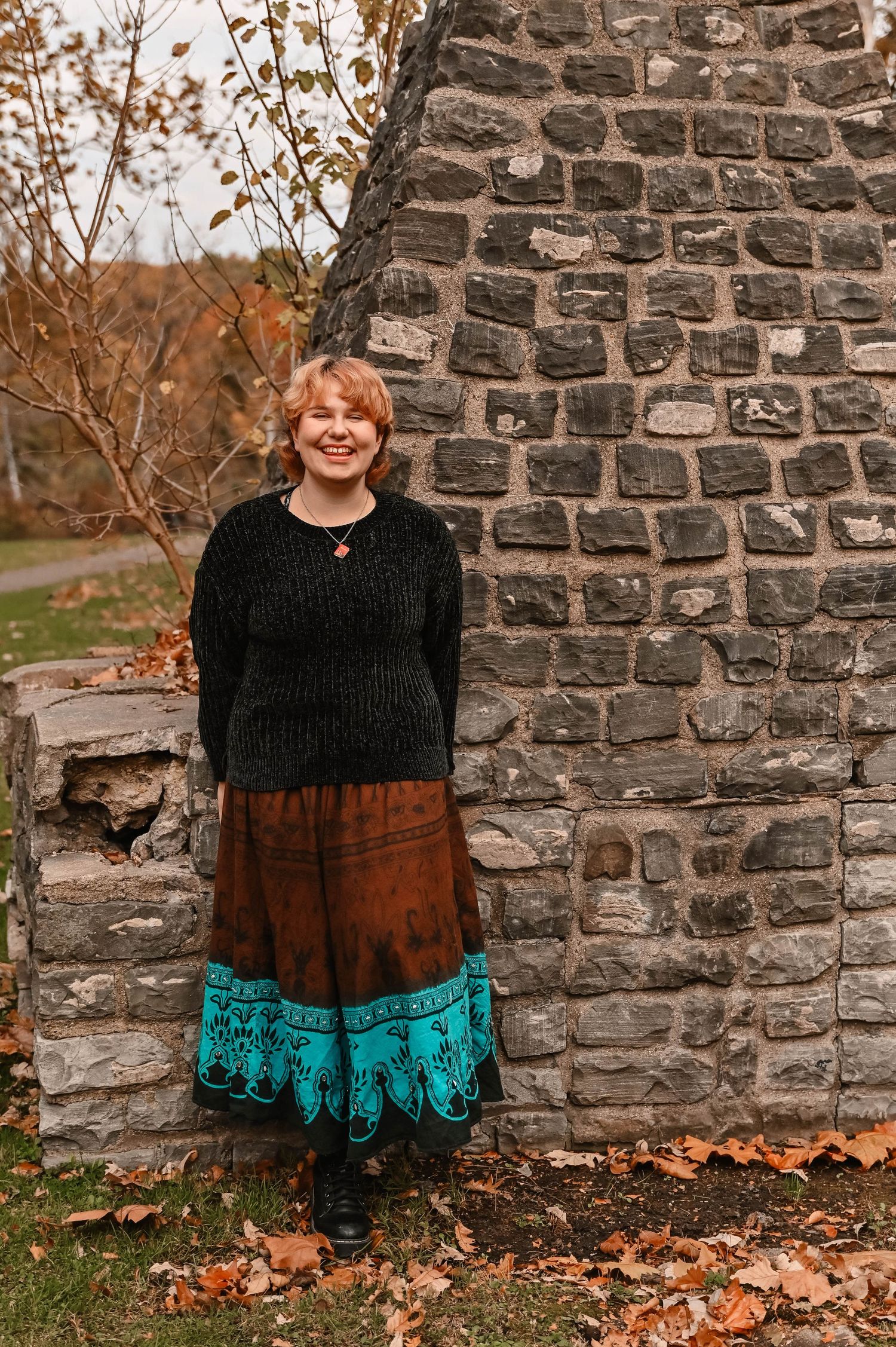 Person in black sweater and colorful skirt posing by stone wall with fallen autumn leaves.
