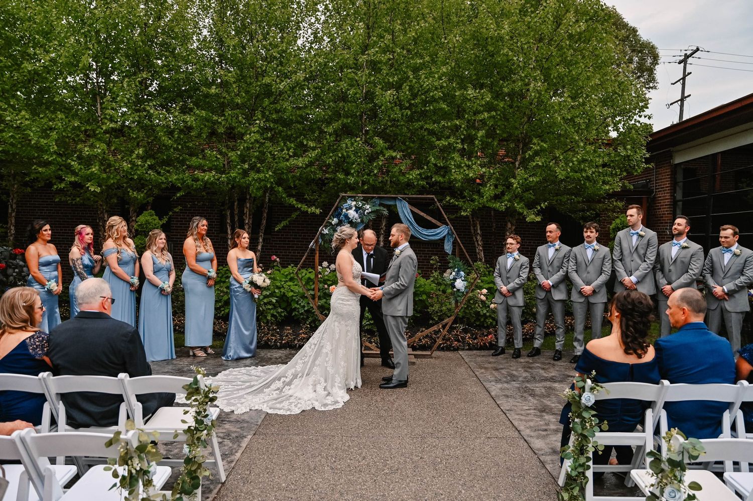 Outdoor wedding ceremony with bridal party in blue dresses and grey suits under trees.