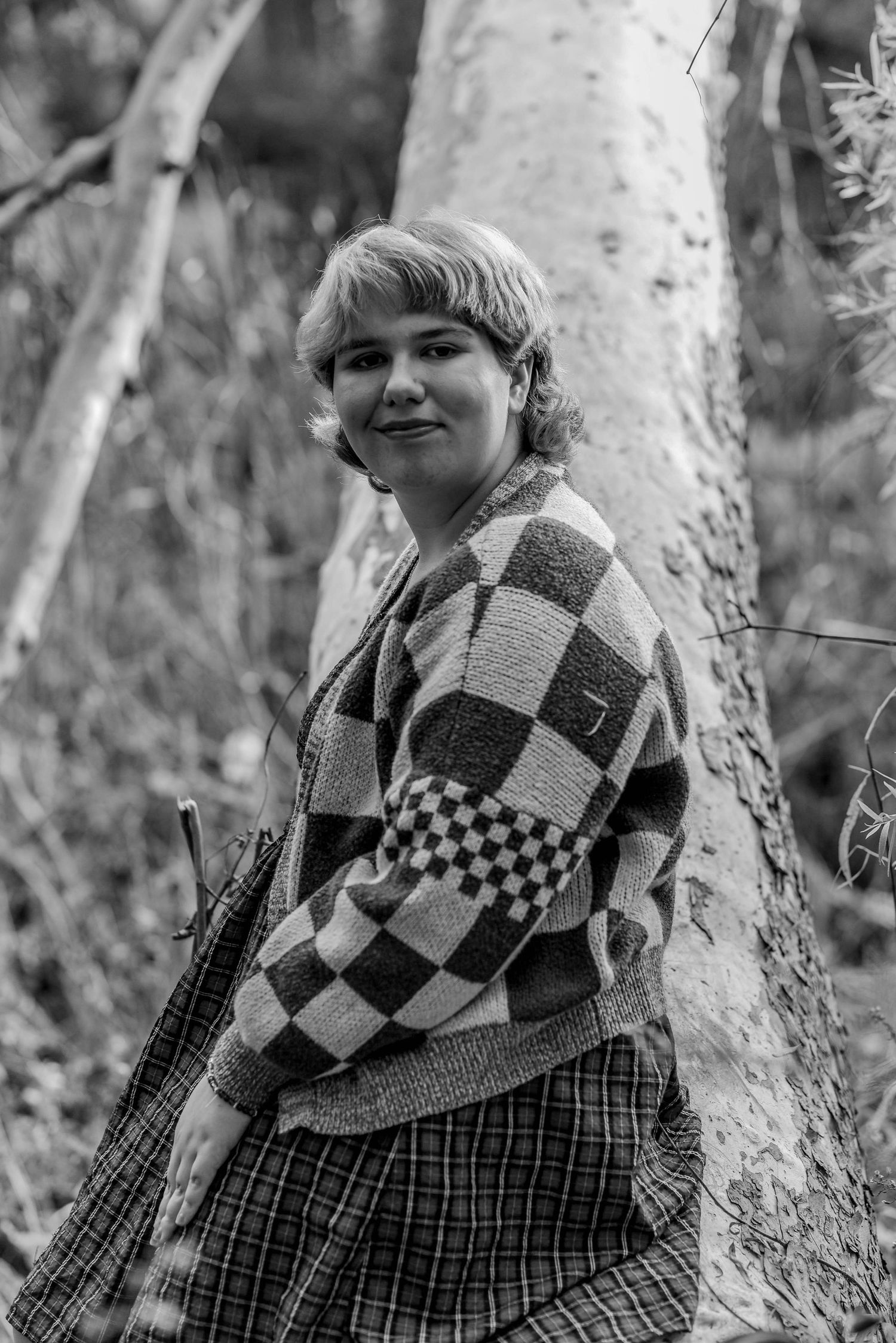 Person wearing a checkered sweater poses against a tree trunk in black and white photography.
