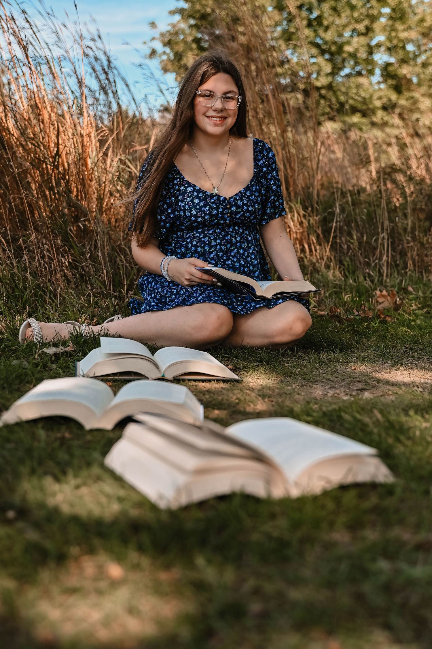 A person in a blue polka dot dress sits outdoors reading a large open book in the grass during golden hour.