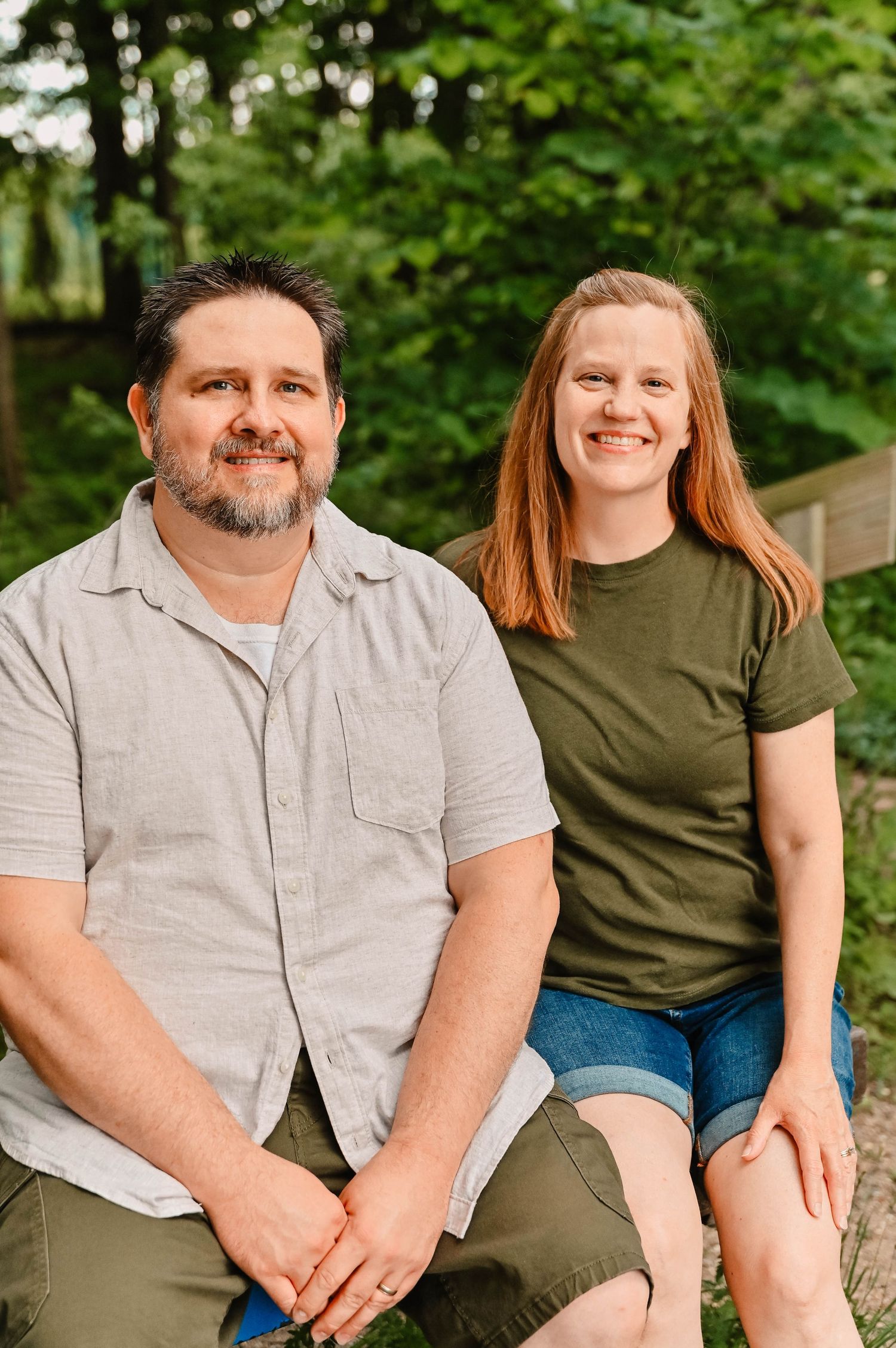 A couple sitting together outdoors in casual attire with green foliage in background.