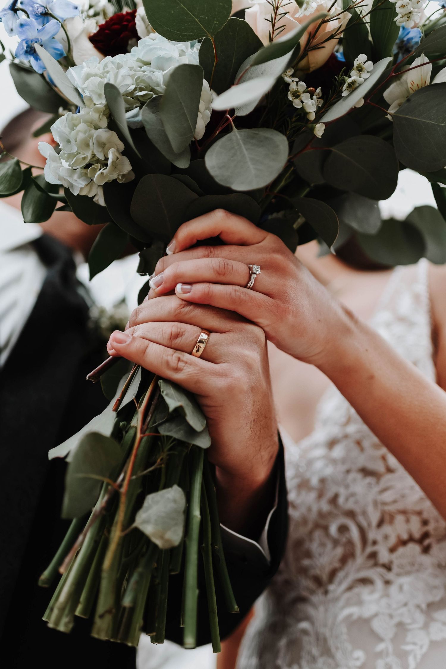 Wedding bouquet with white flowers and eucalyptus leaves held by hands showing rings.