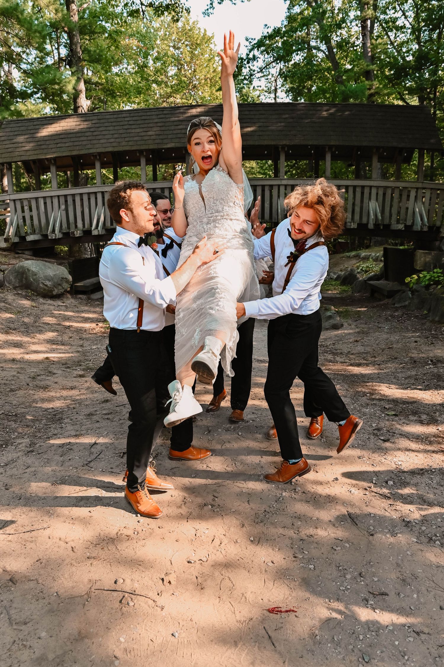 Wedding party members lift someone in white dress for fun photo at outdoor venue.