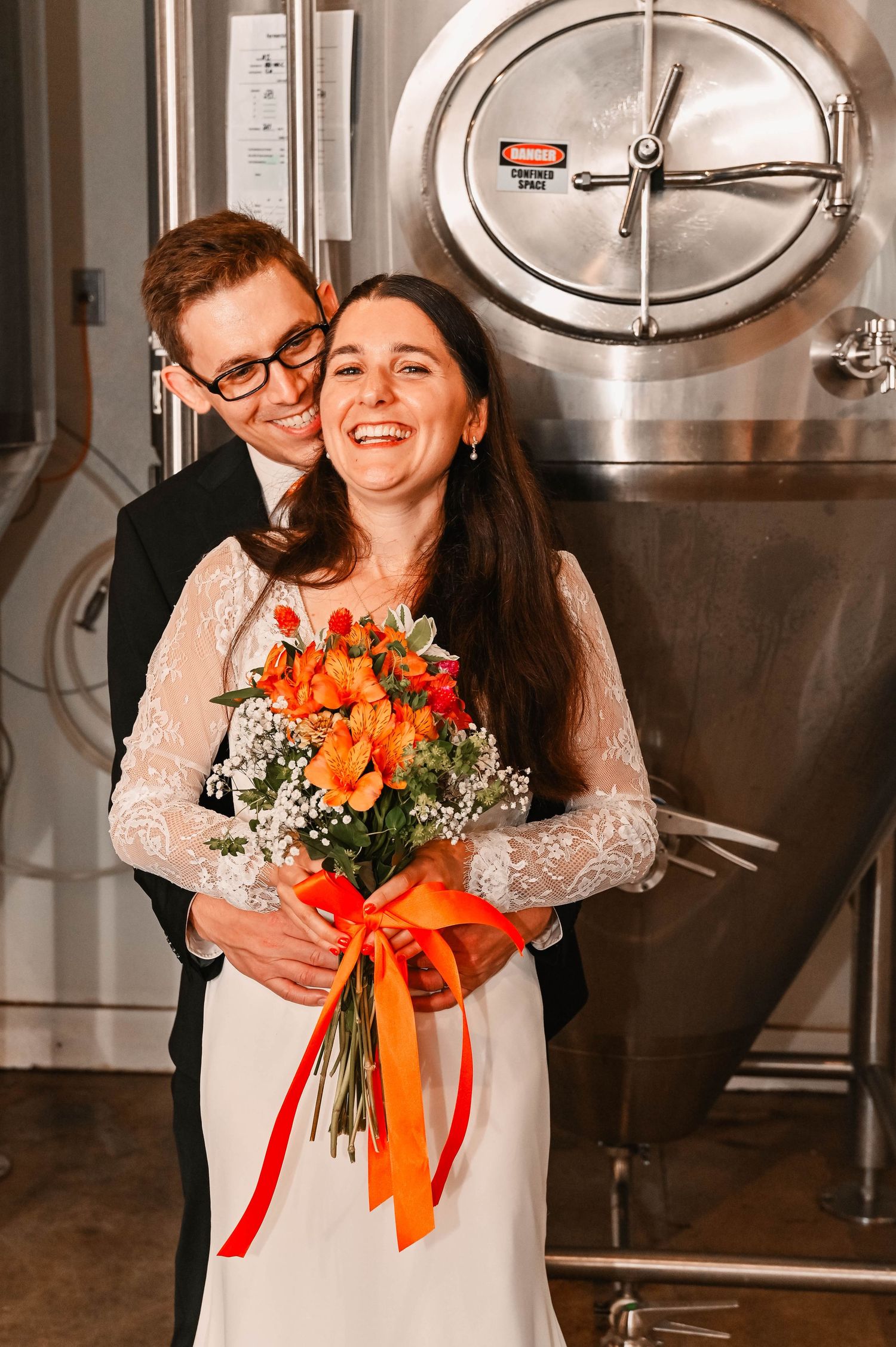 Couple laughs while holding orange and white floral bouquet in industrial brewery setting.