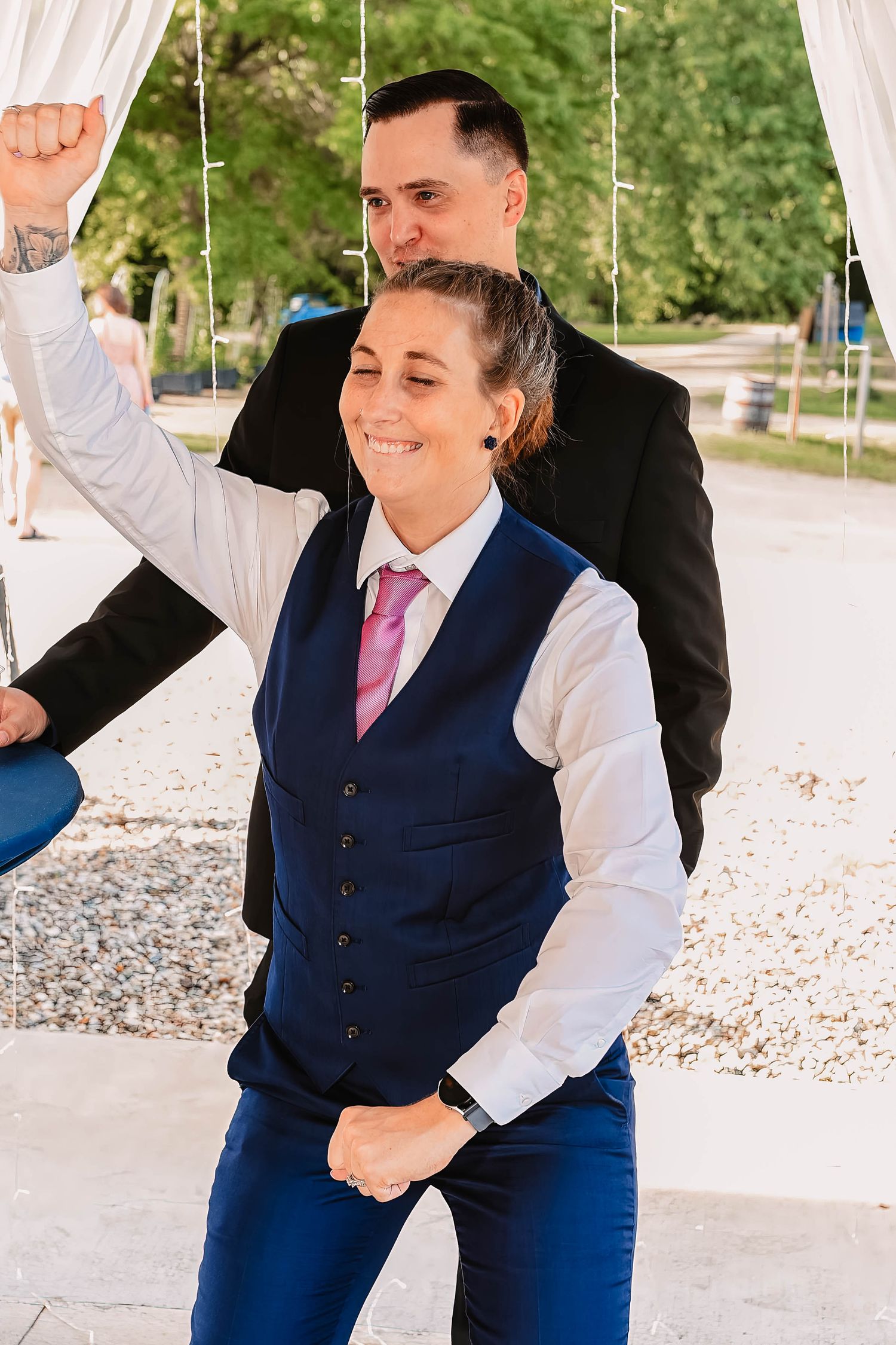 Wedding party members in navy vests celebrate with raised arms under white draping.