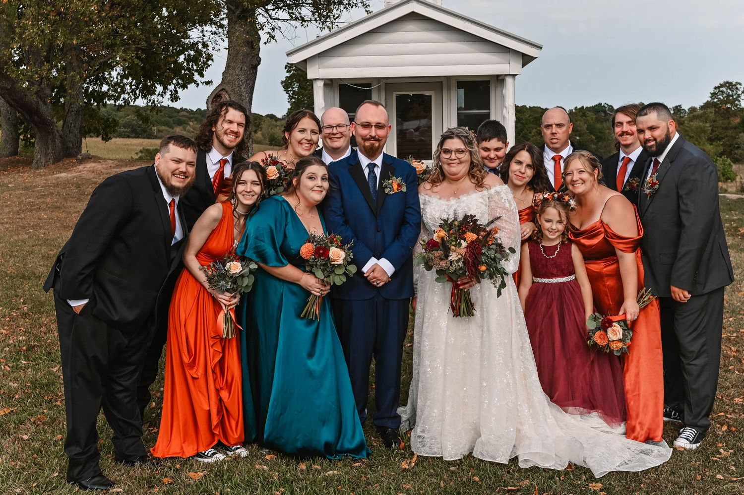 Wedding party in autumn colors poses outdoors with bridesmaids in burnt orange and teal dresses.