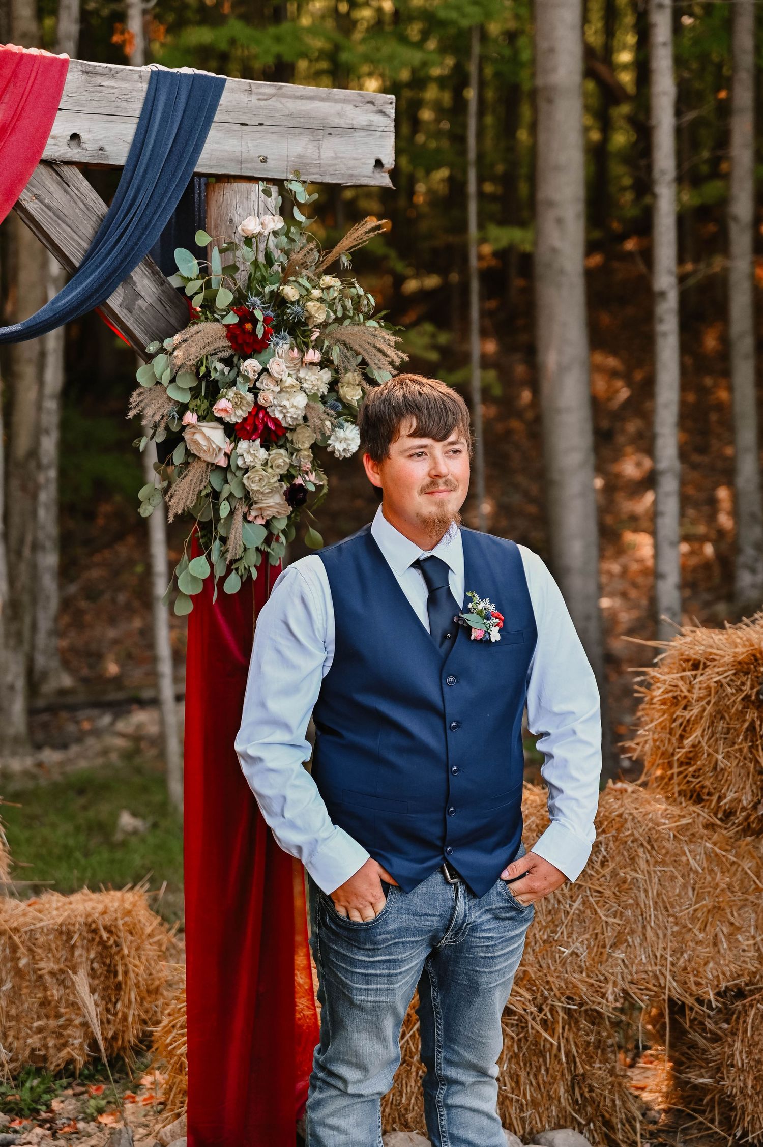 A young attendant in a navy vest and light blue shirt poses by hay bales at an outdoor wedding.