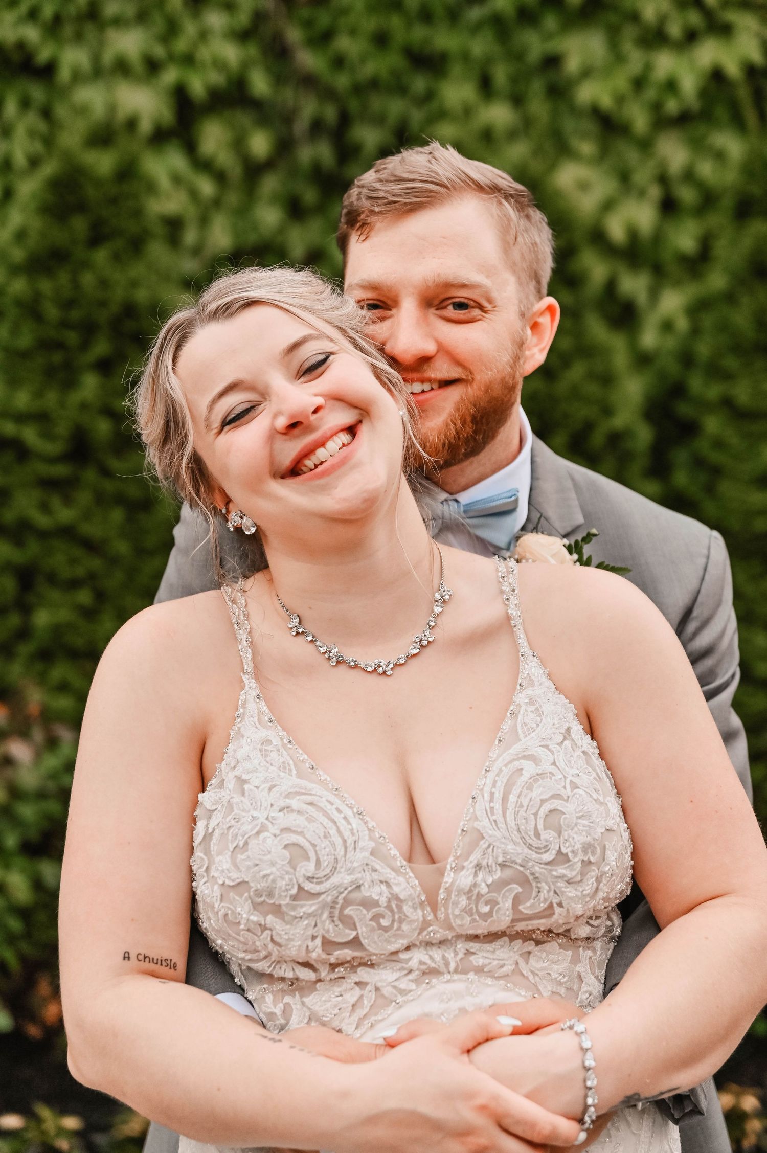 A couple shares an intimate moment in formal attire against a green hedge background.