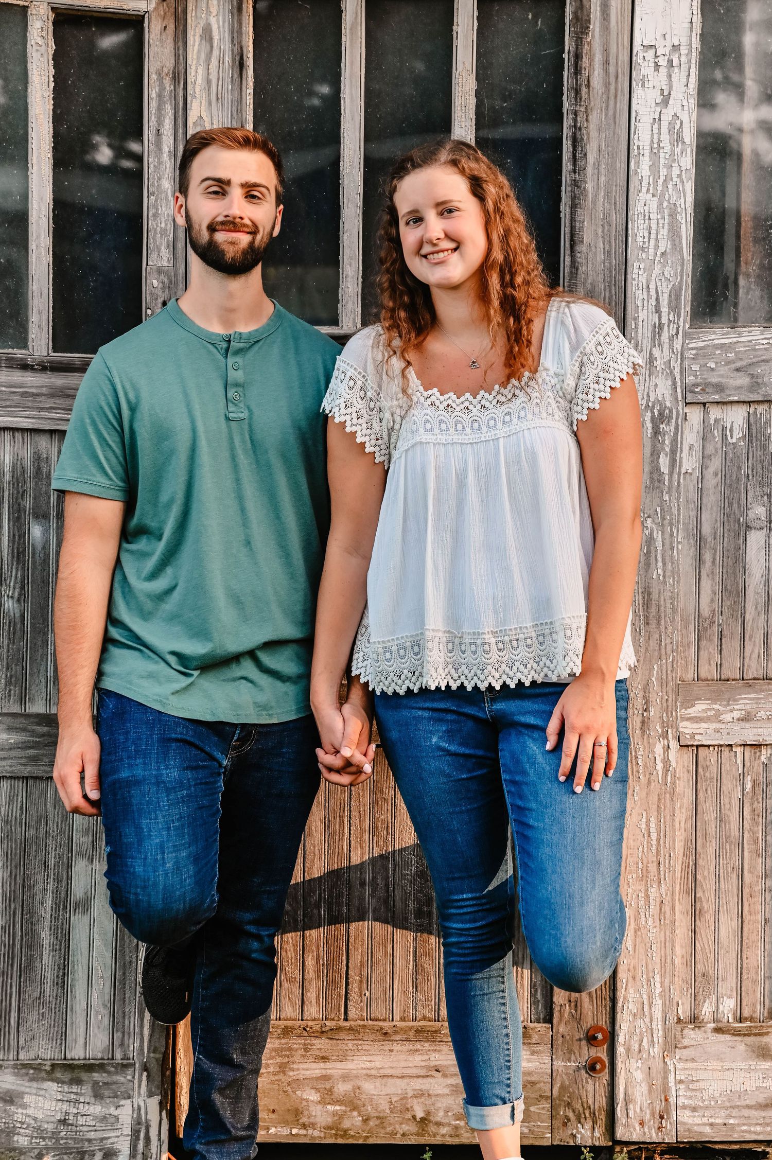 A couple stands together against a rustic wooden wall wearing casual outfits.