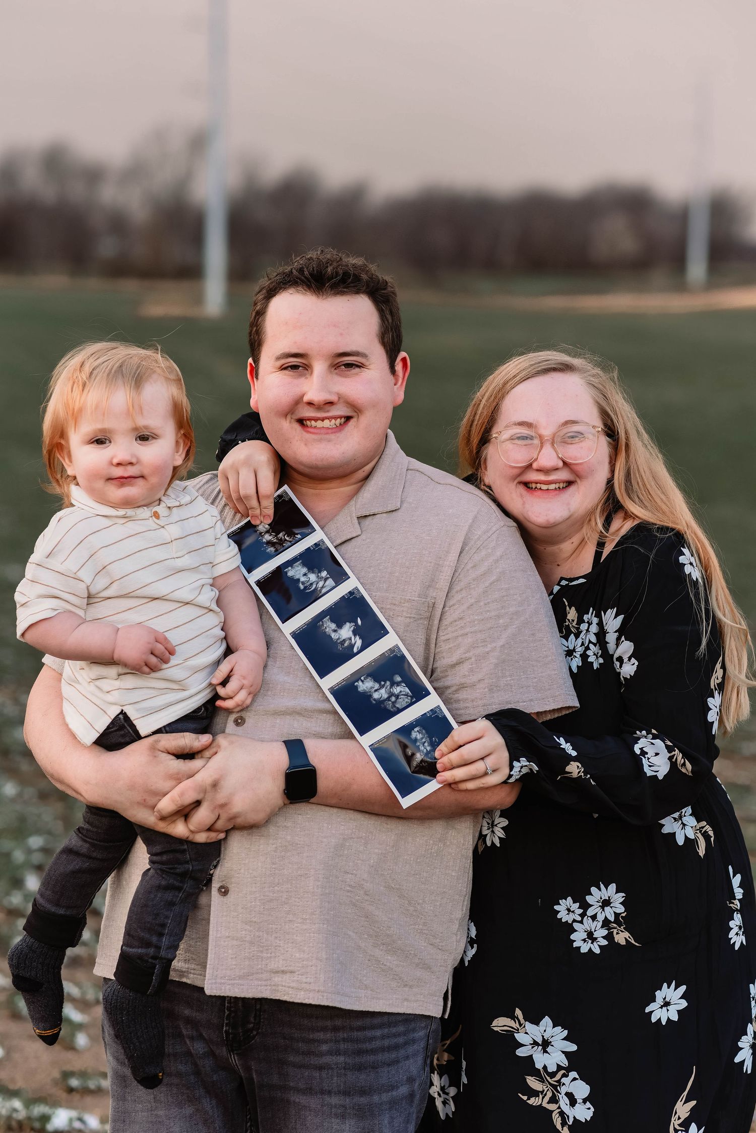 Family holds ultrasound photos while posing together outdoors for pregnancy announcement.