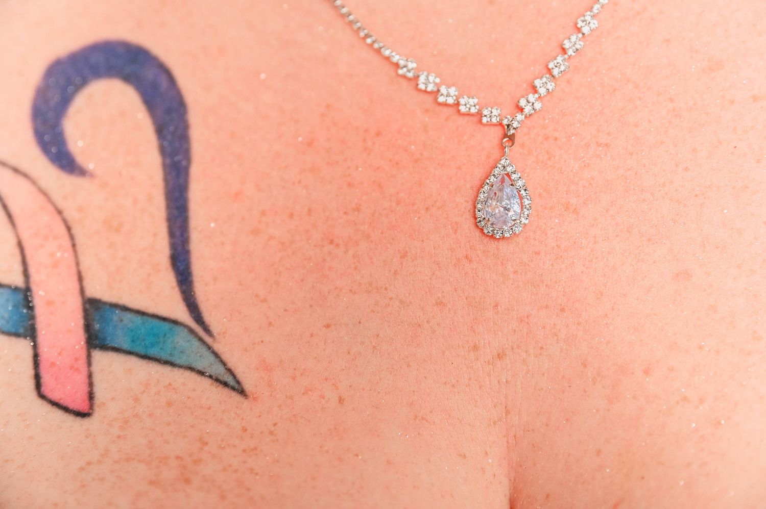 Close-up of a blue ribbon tattoo and crystal necklace on skin.
