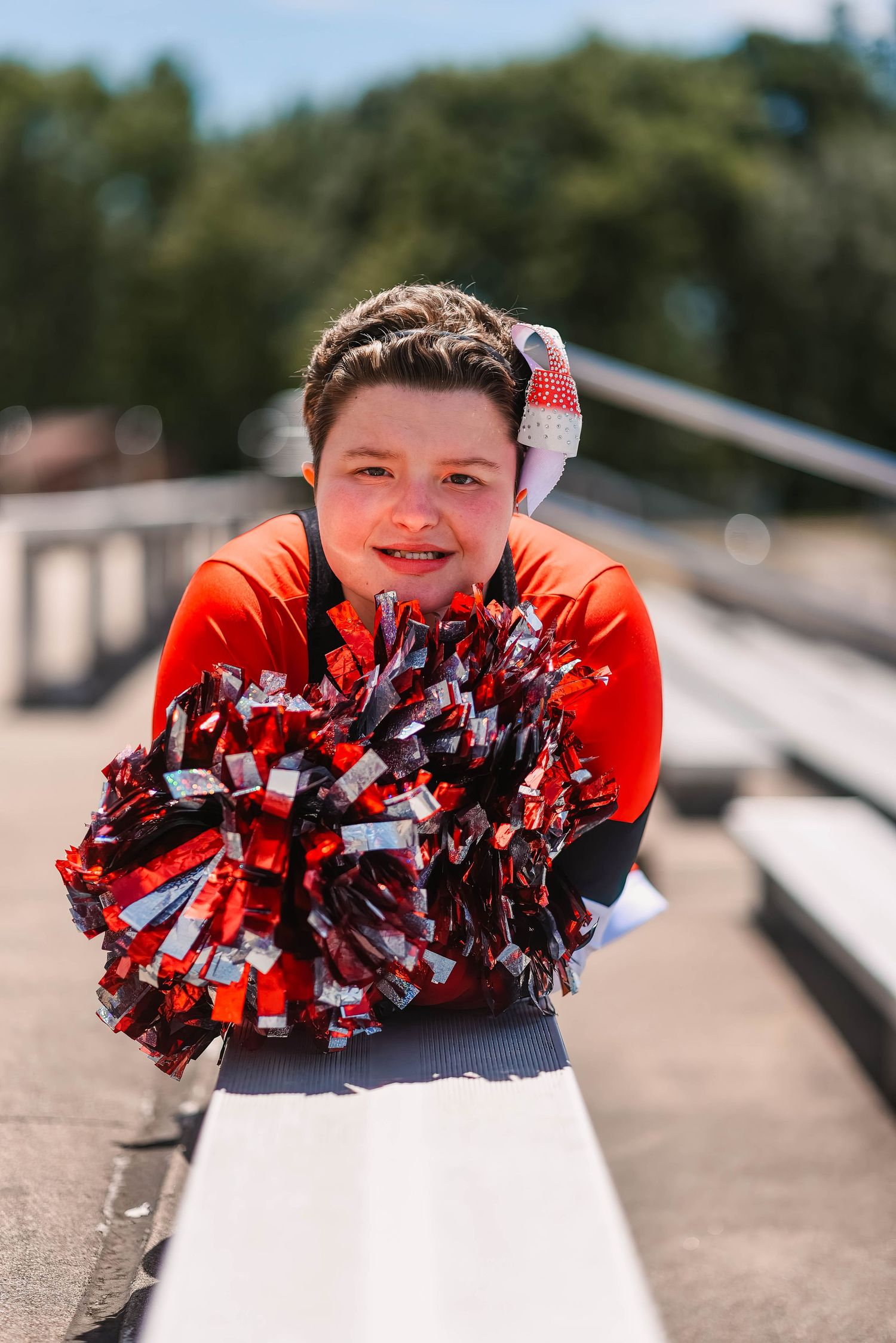 Cheerful student in orange shirt holds red and white pom poms on bleachers.