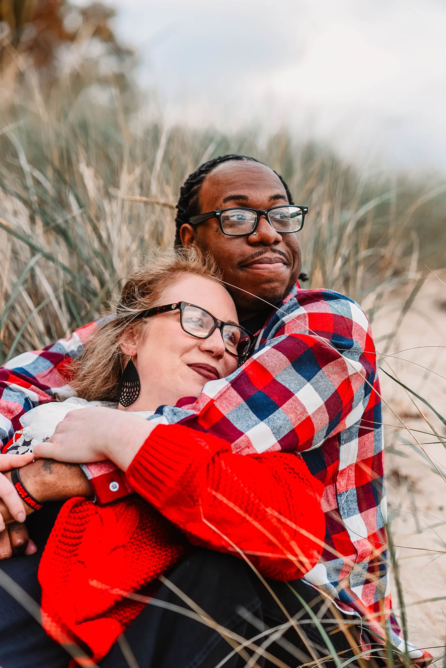 A couple in casual plaid shirts embrace among beach grass on a cloudy day.