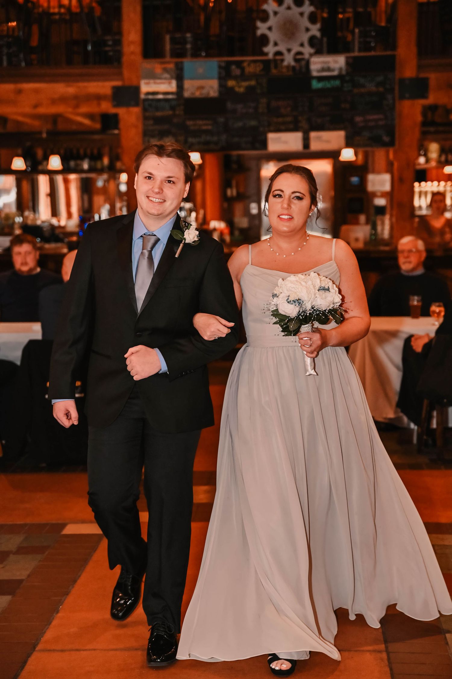Wedding party members walk arm in arm down a dimly lit venue aisle wearing formal attire.