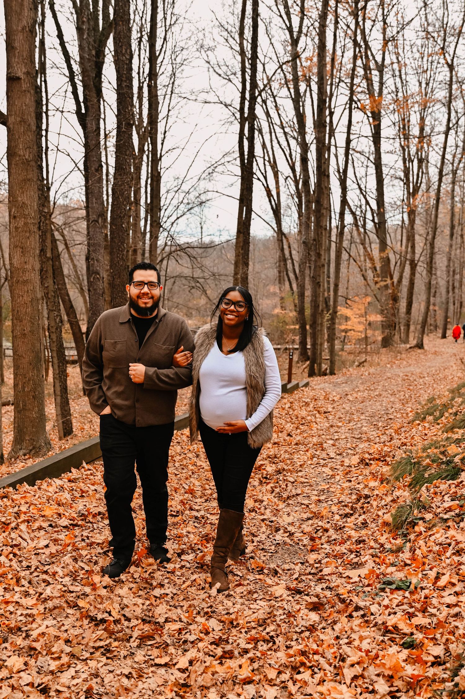 A couple walks through fallen autumn leaves in a serene forest setting.