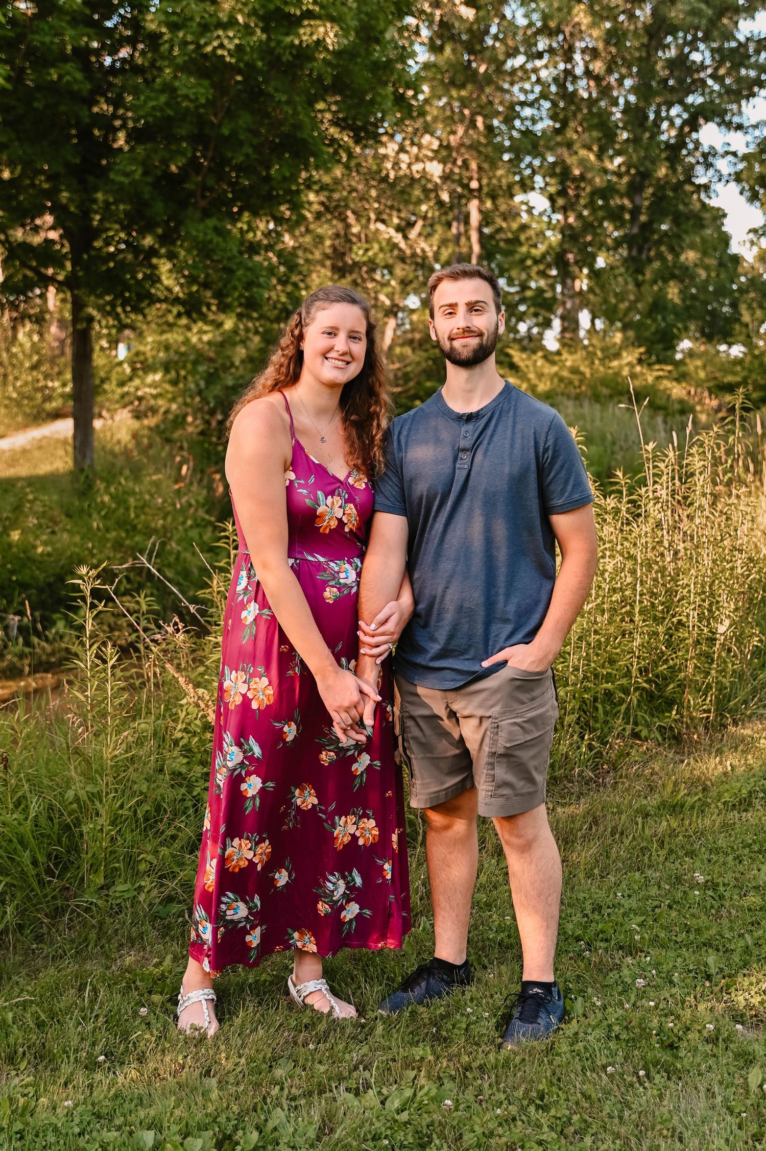 Couple poses together in summer field with golden sunlight and trees.