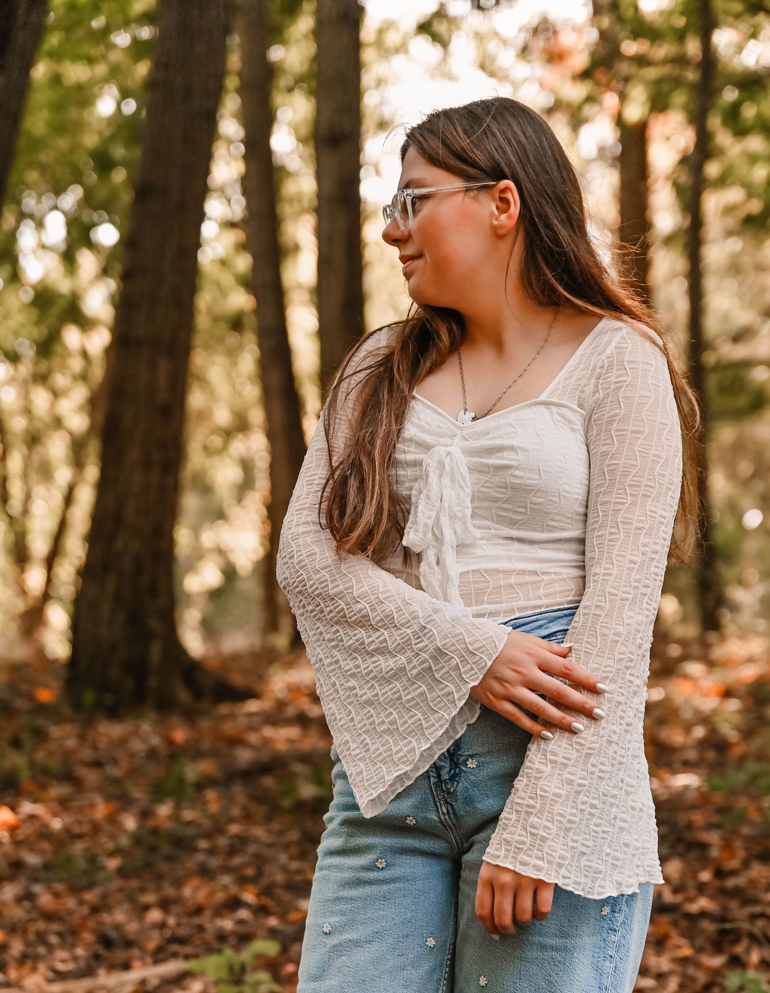Someone in white lace top and denim poses casually against autumn forest backdrop.