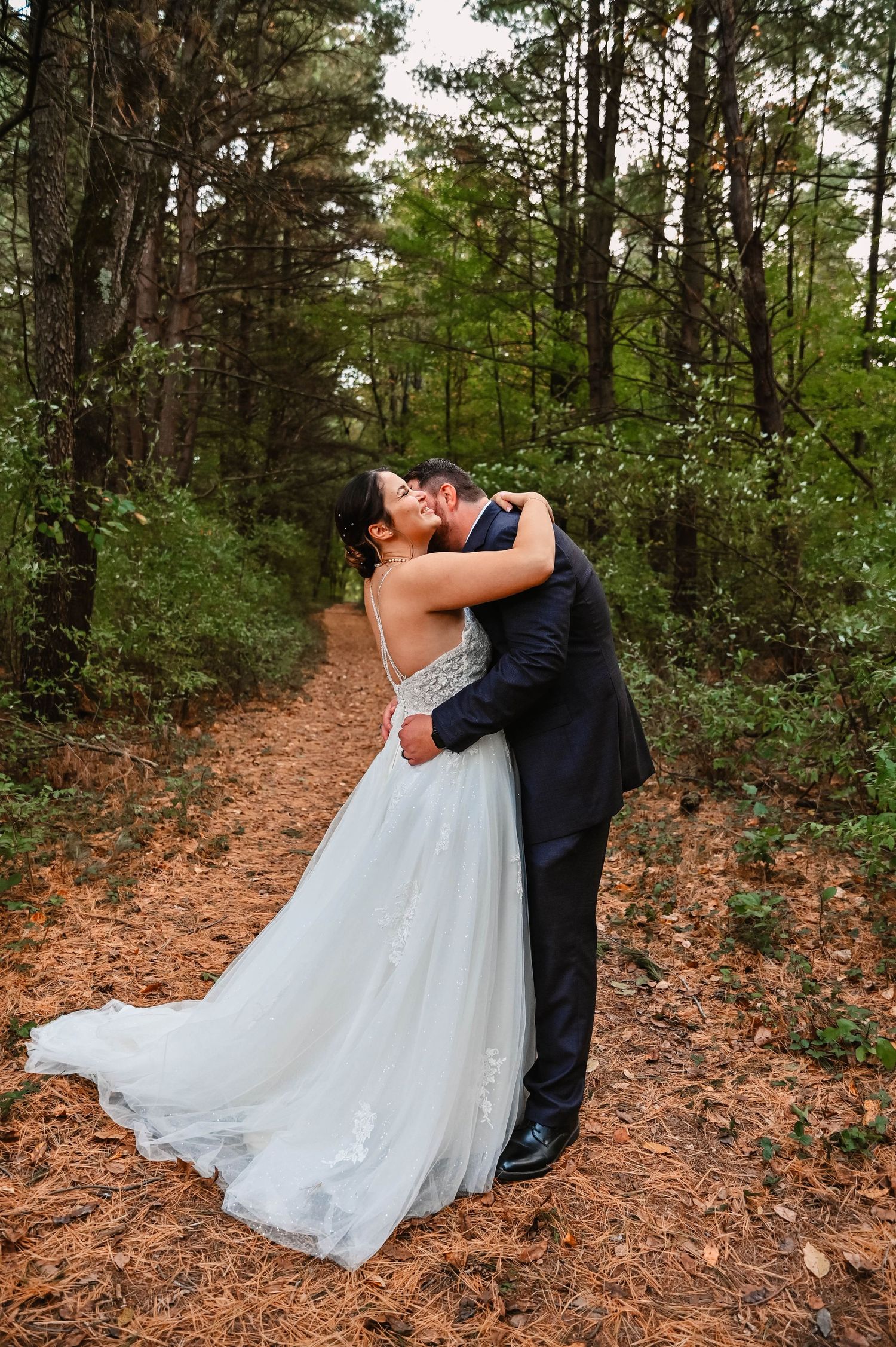 A romantic wedding couple embraces on a forest path surrounded by tall pine trees.
