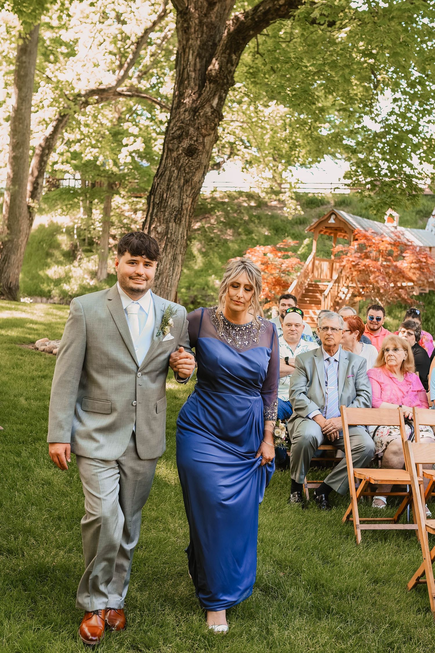 Wedding ceremony guests seated on wooden chairs under large trees in outdoor garden setting.