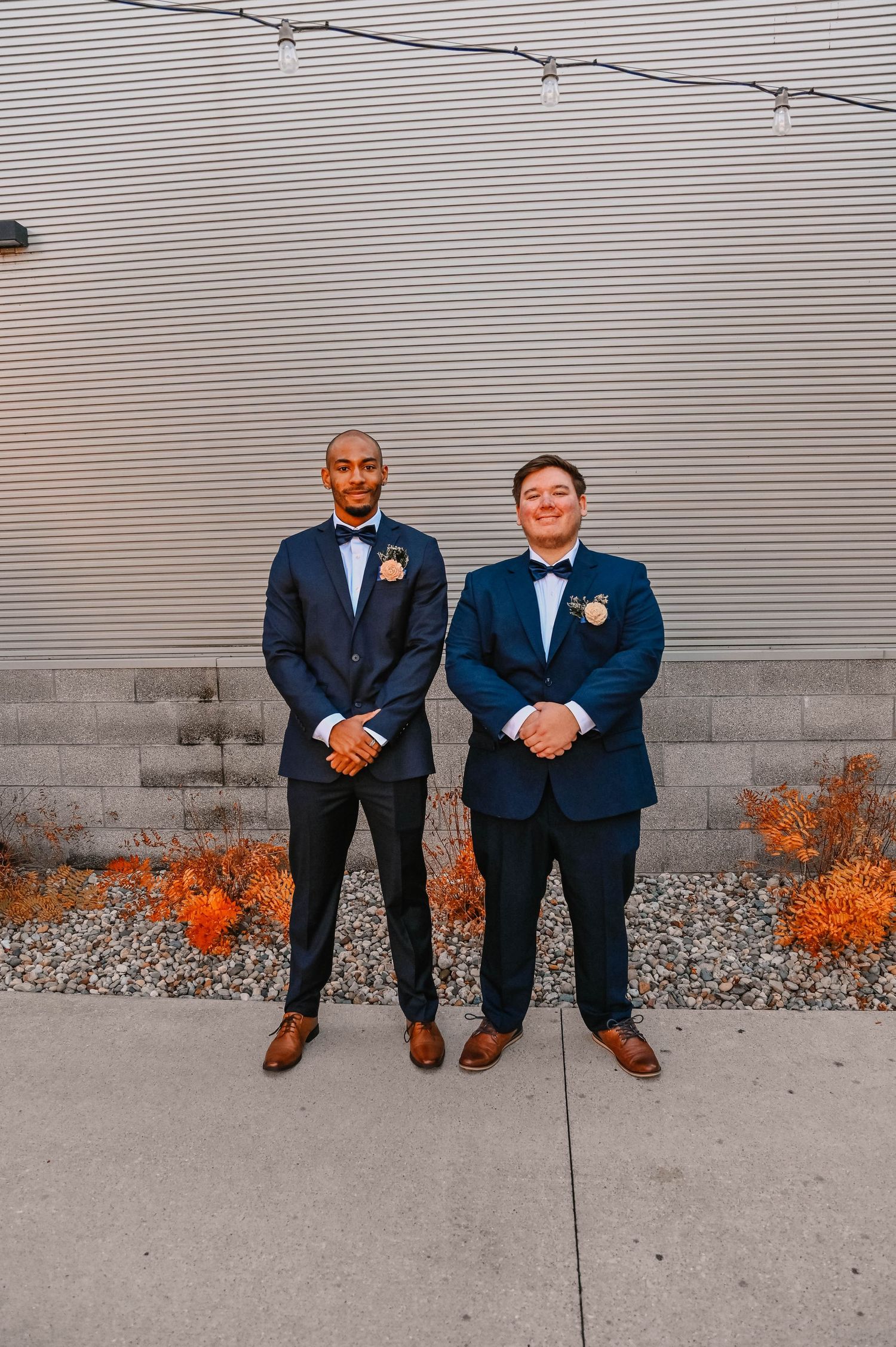 Two groomsmen in navy suits and bow ties pose against a grey industrial wall with orange accents.