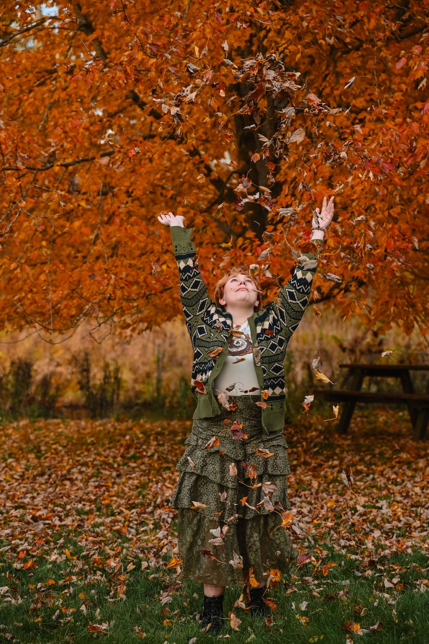 Person joyfully tosses orange fall leaves in the air under autumn trees.