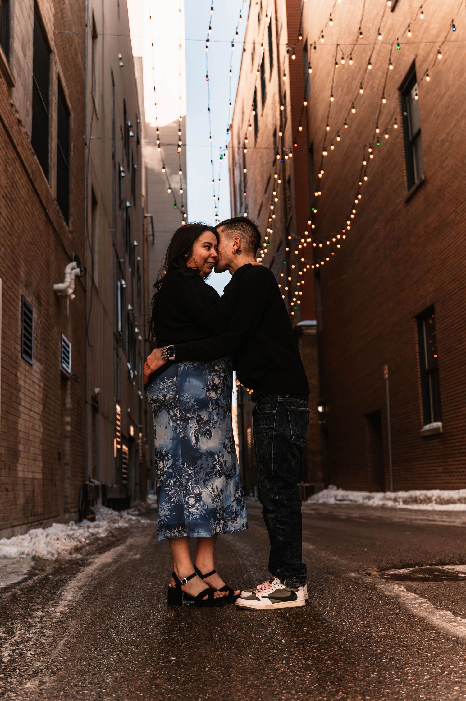 String lights illuminate a romantic moment in a brick alleyway covered in snow.