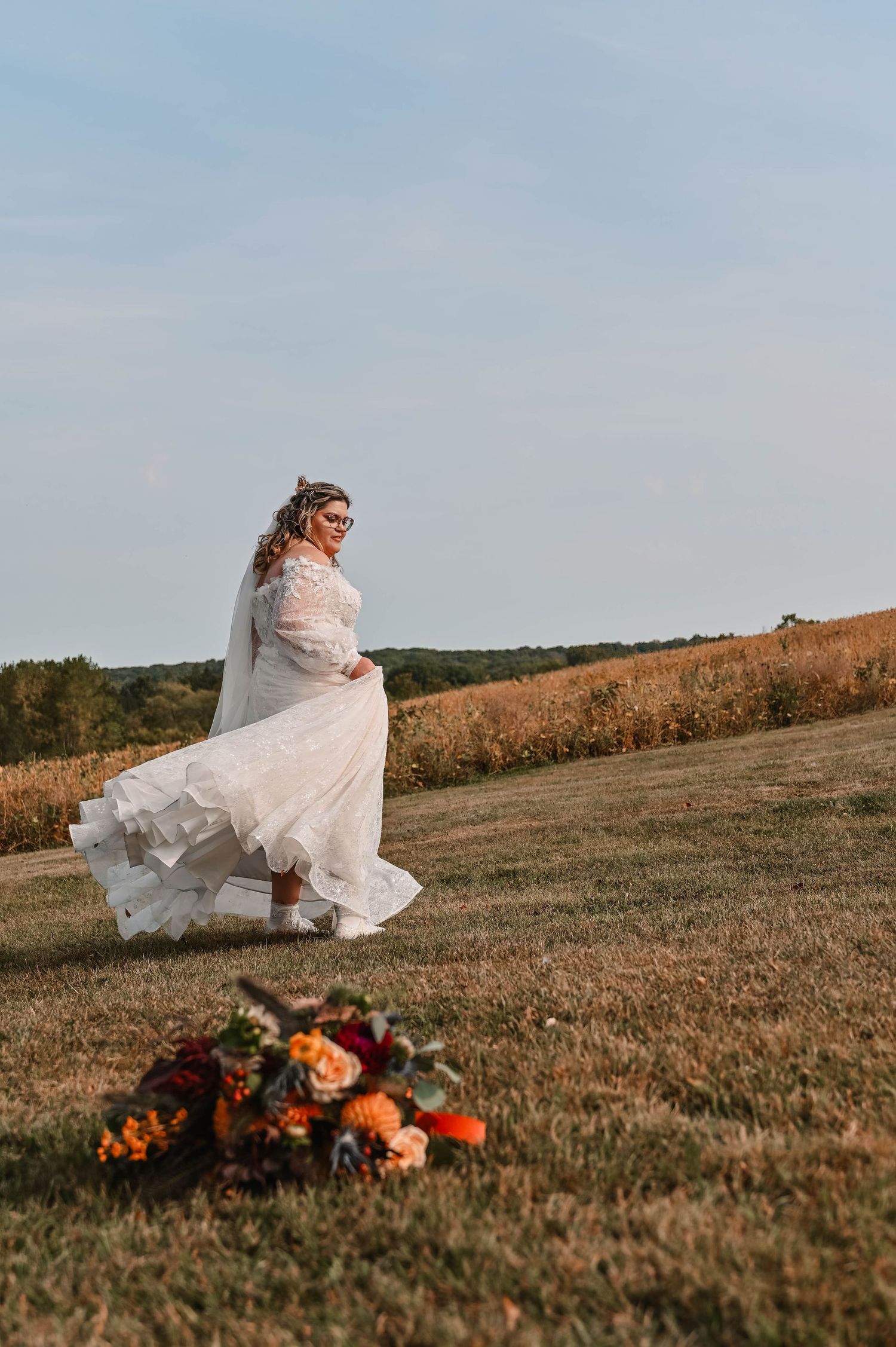 Bride in white dress posing with orange floral arrangement in an autumn field at sunset.