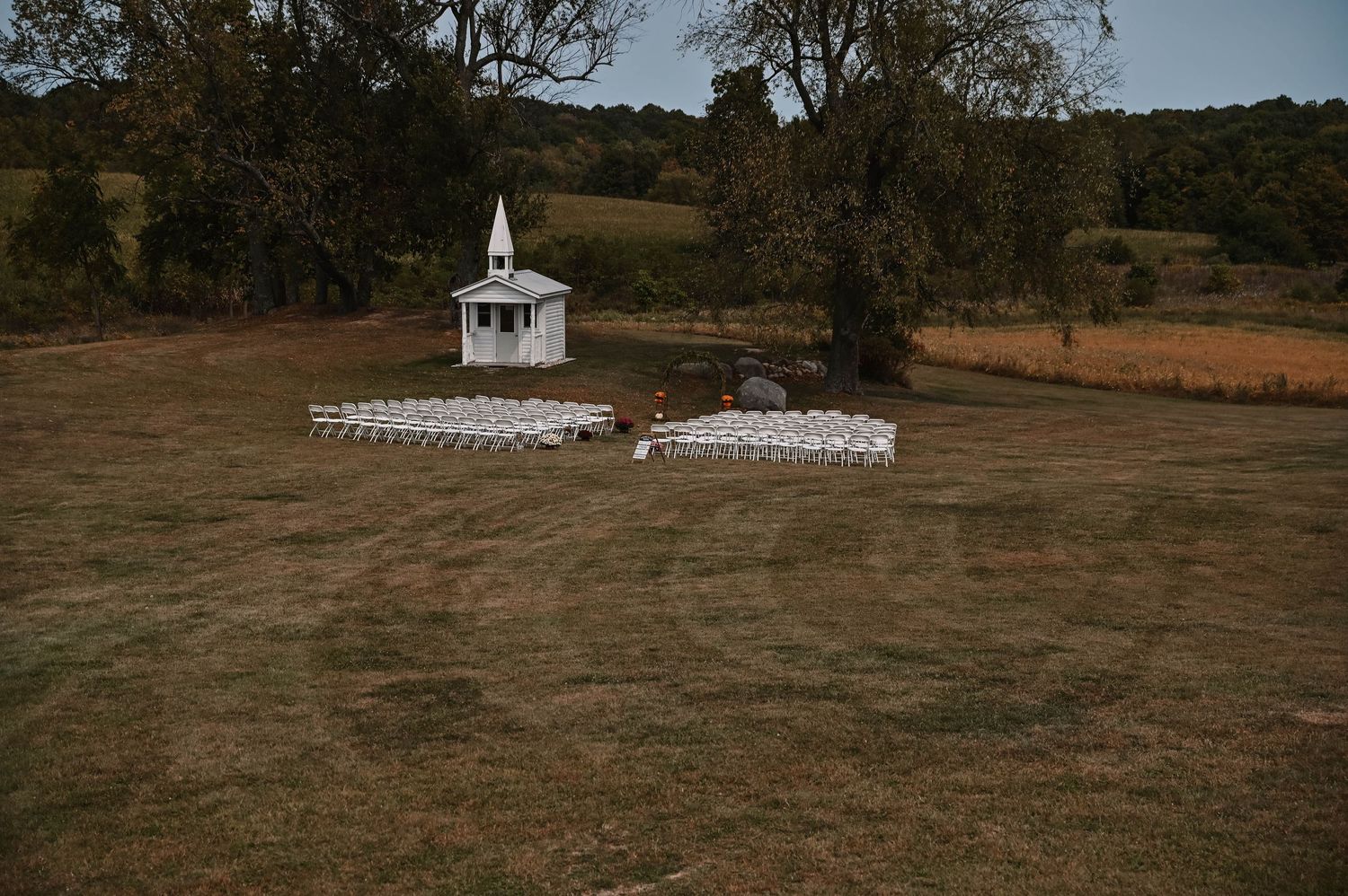 Outdoor wedding venue setup with white chairs and a gazebo in a rustic field setting.