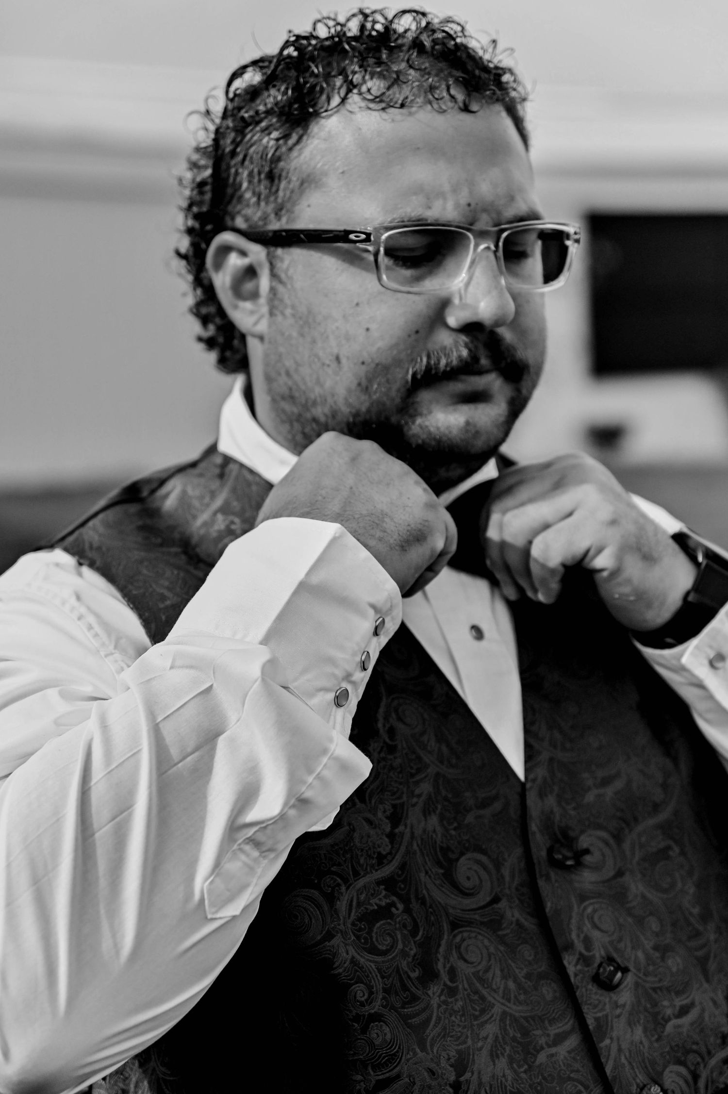 Black and white portrait of a groom adjusting his tie while wearing a patterned vest and glasses.
