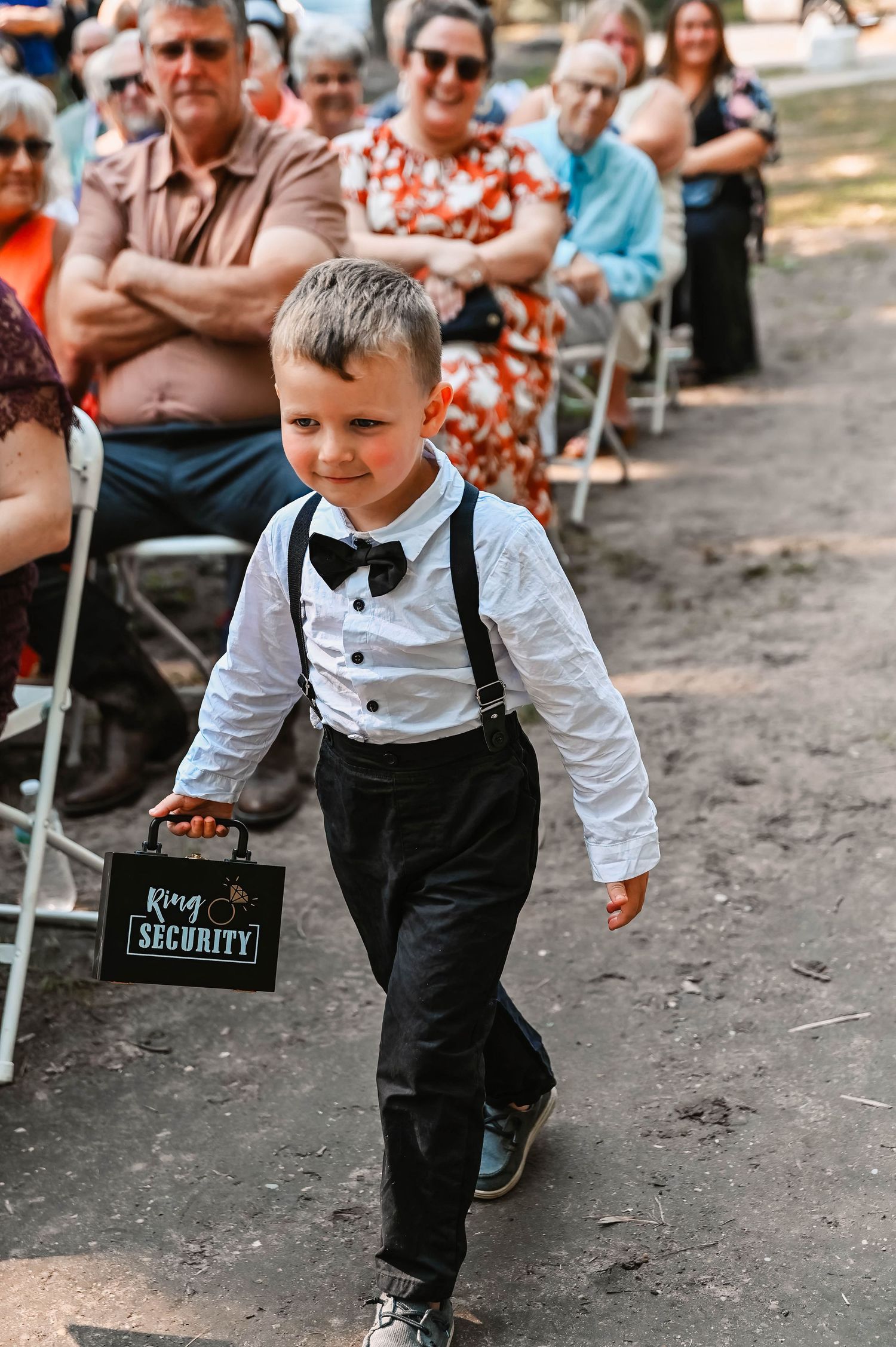 Young ring bearer in suspenders and bow tie carries a security sign at an outdoor wedding ceremony.