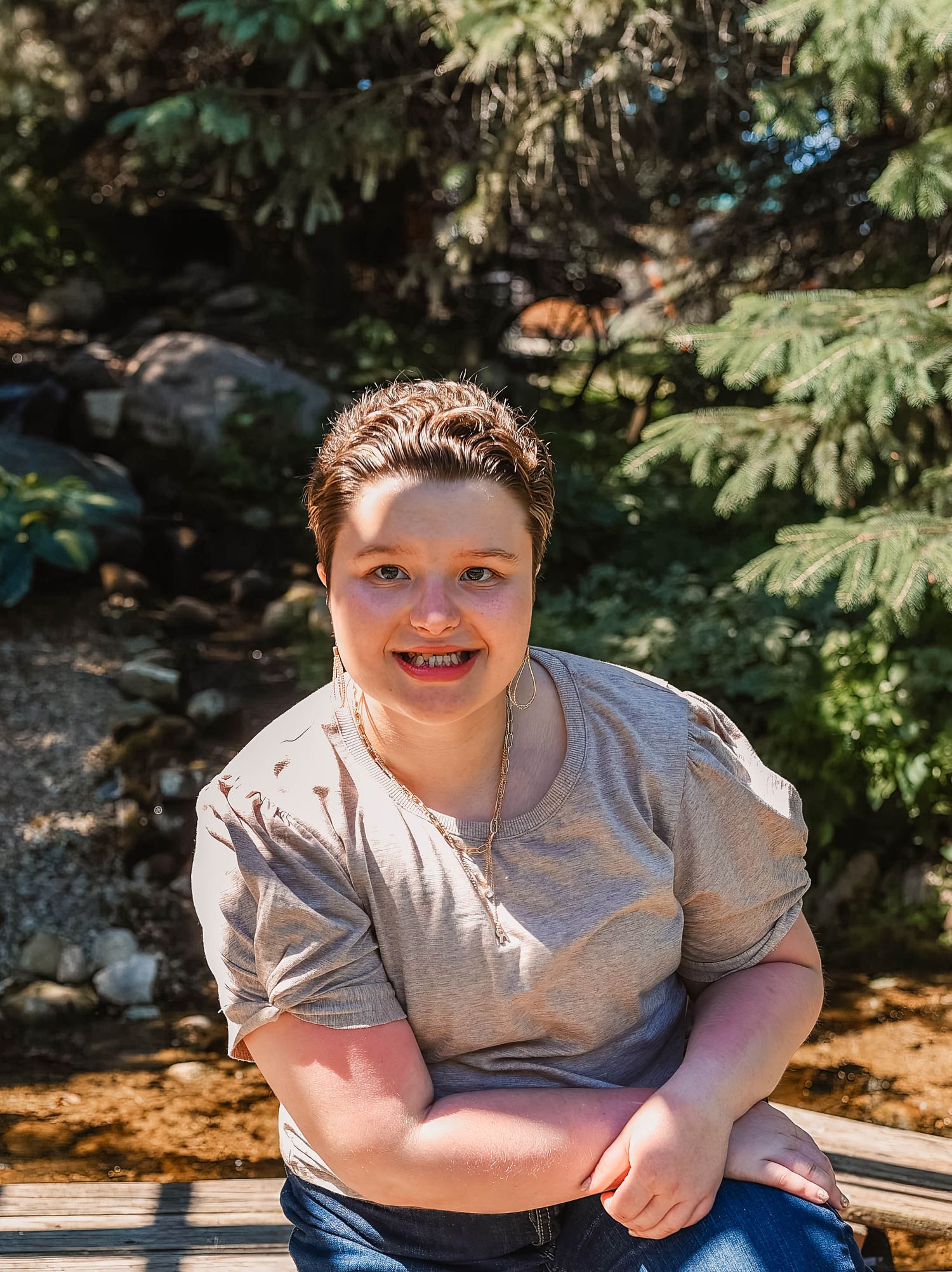 A person in a gray t-shirt sits outdoors in a wooded area with dappled sunlight filtering through the trees.